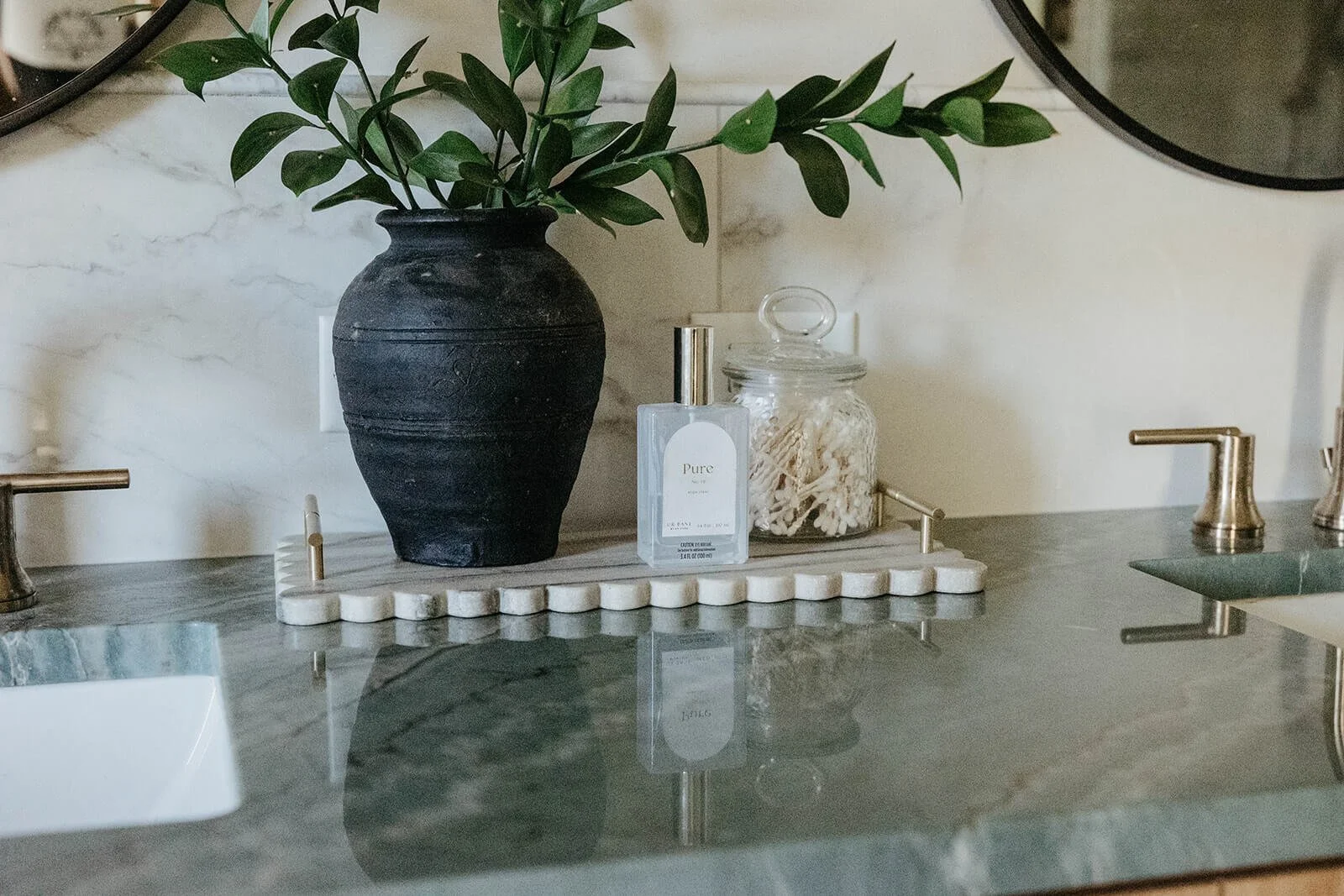Bathroom countertop close-up showing a dark green marble or stone counter, a black vase with greenery, and a selection of soaps and cotton balls on a tray between two gold faucets.