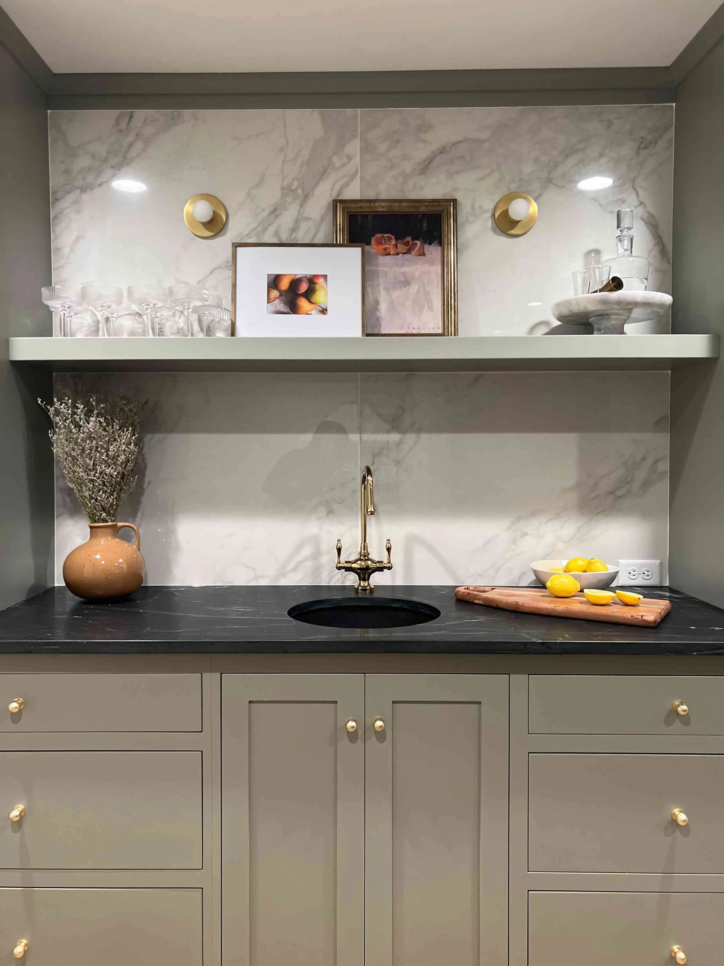 Close-up of the wet bar sink and shelving with a dramatic marble backsplash. The area features gold sconces, framed art, and a matte brass faucet.