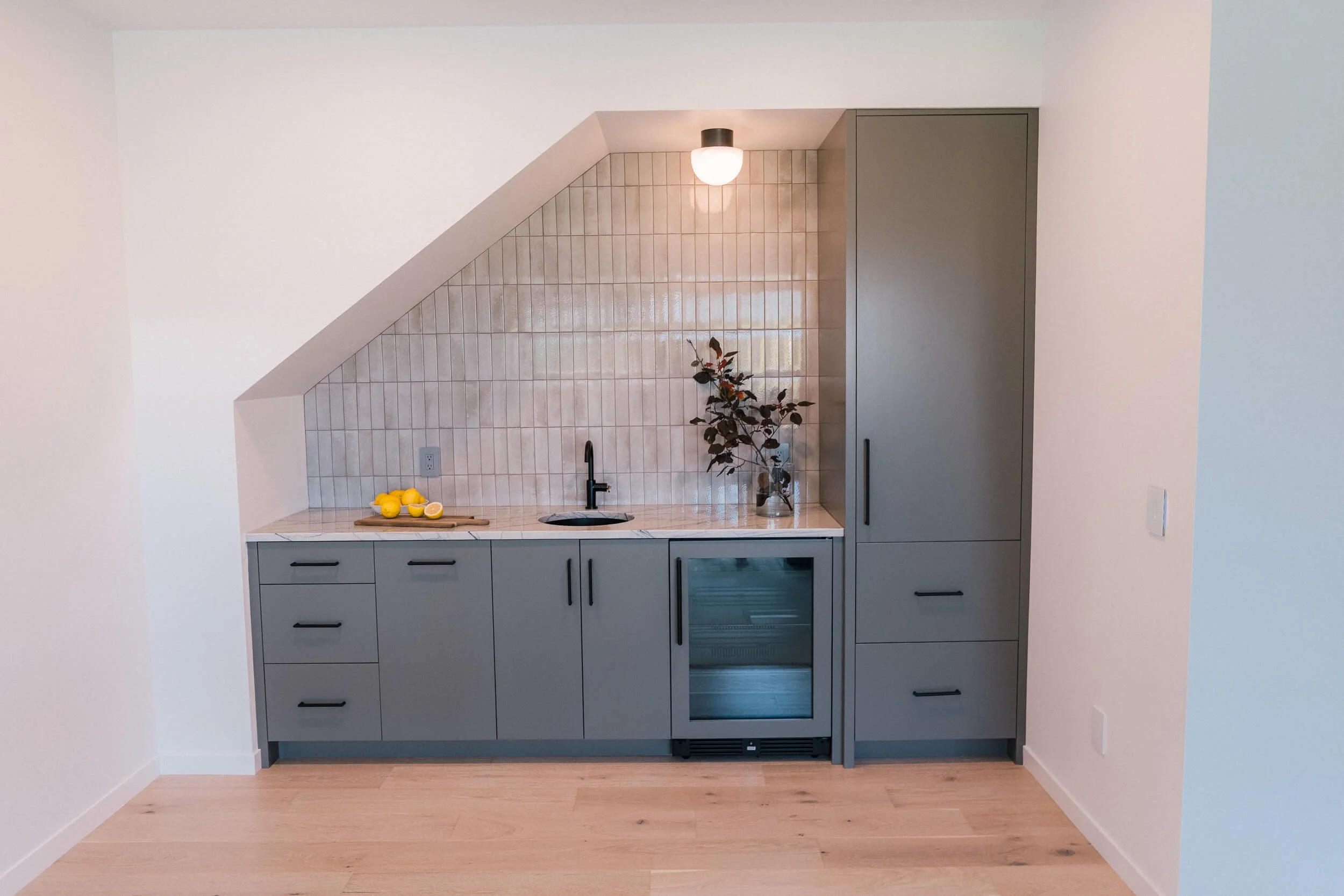 Small under-stair wet bar with dark gray flat-panel cabinets, light wood counter, a tiled backsplash, and under-counter refrigerator.