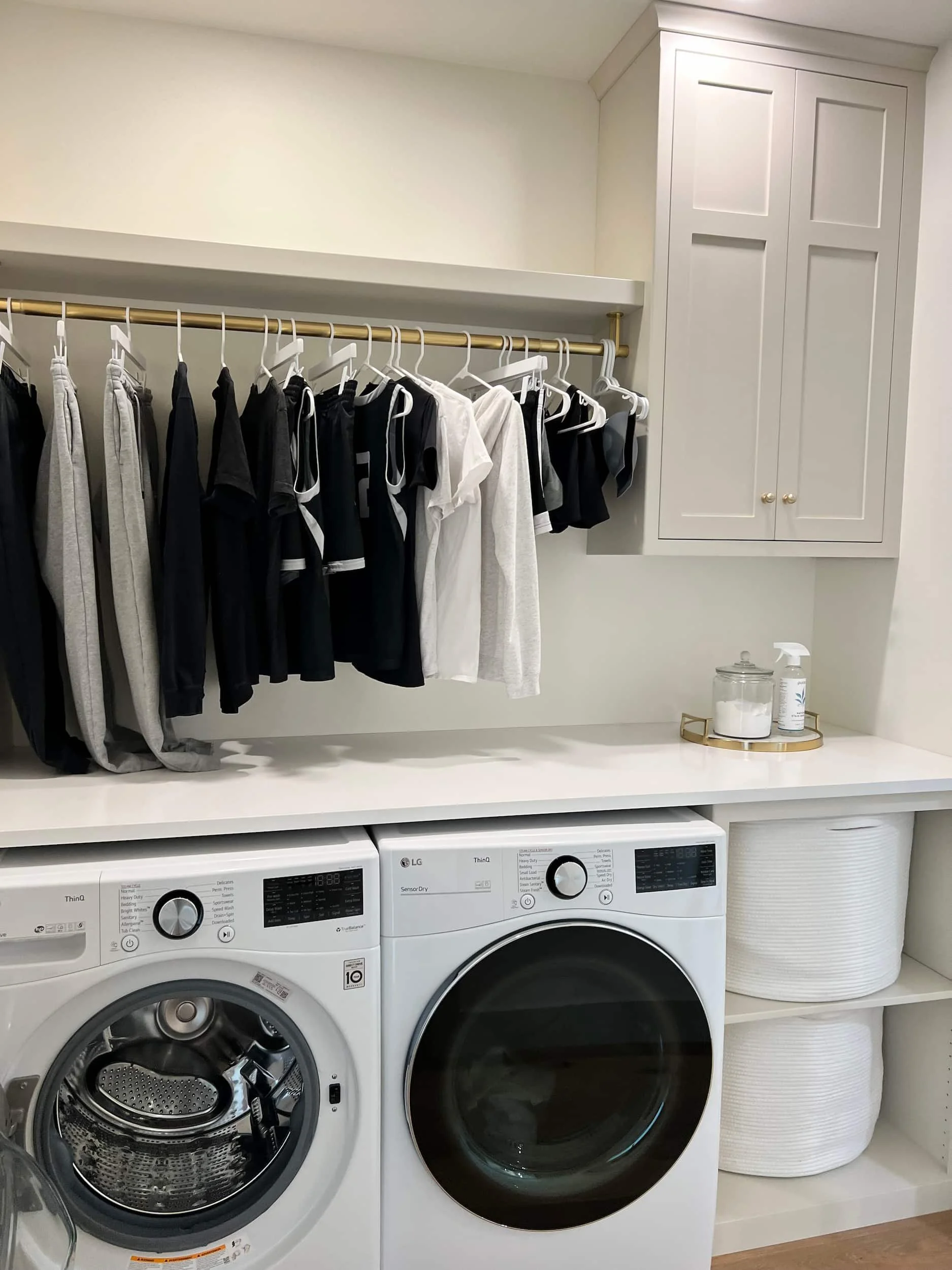 Functional laundry room with stacked washer and dryer units, a white countertop for folding, hanging clothes storage, and closed white cabinets above.
