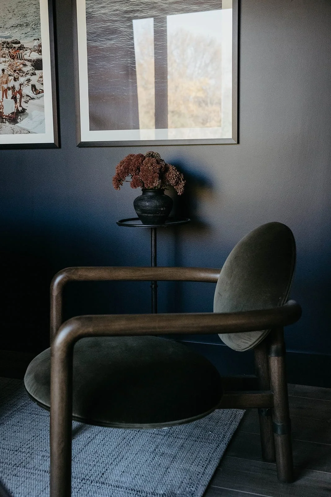 Moody close-up of a modern office armchair with a dark wood frame and a dark velvet cushion, set against a deep blue wall with framed artwork.