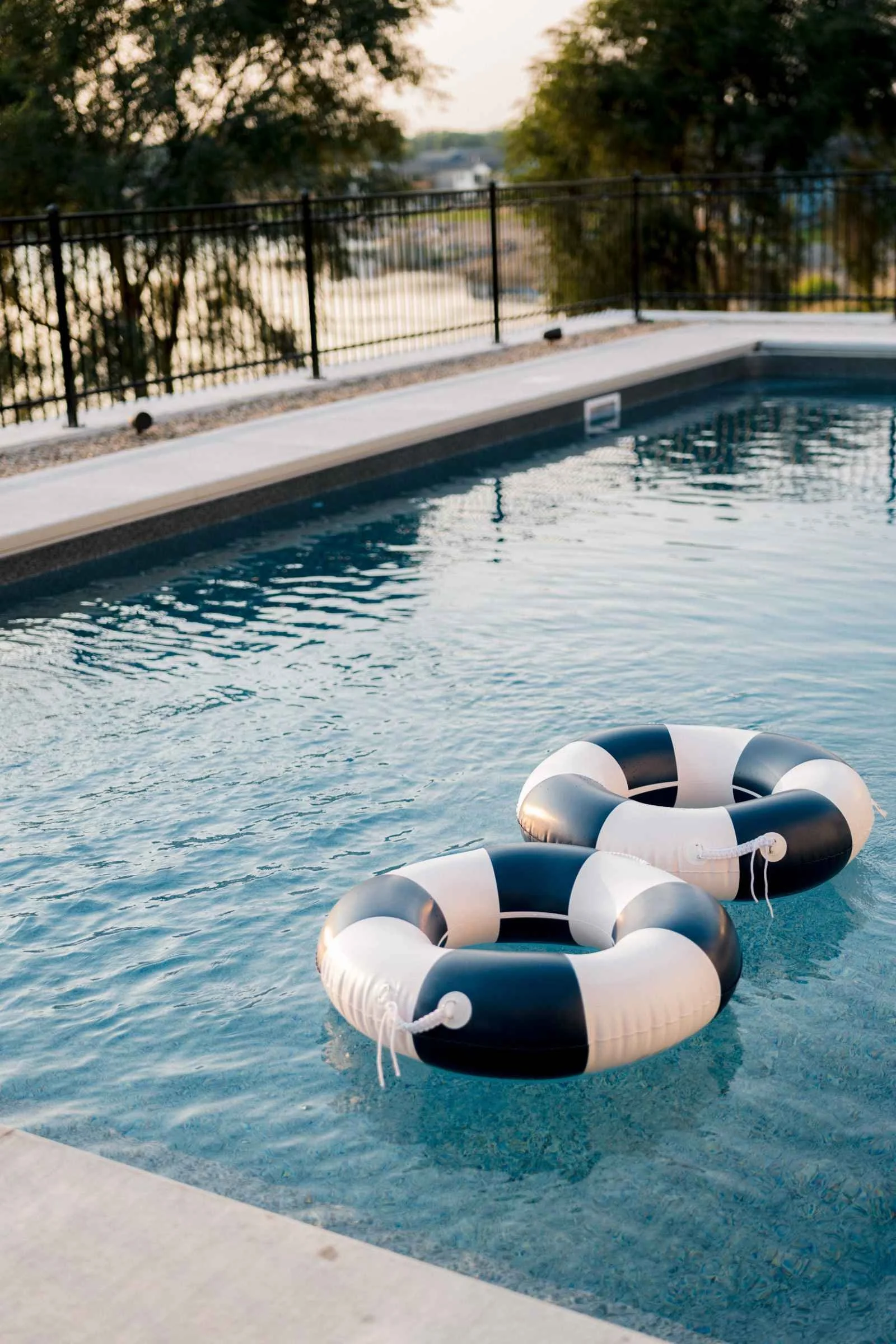 Close-up of the pool water and edge with a view through the black metal fence to the water and trees beyond. Two large navy and white striped ring floats resting on the clear blue water of an in-ground swimming pool.