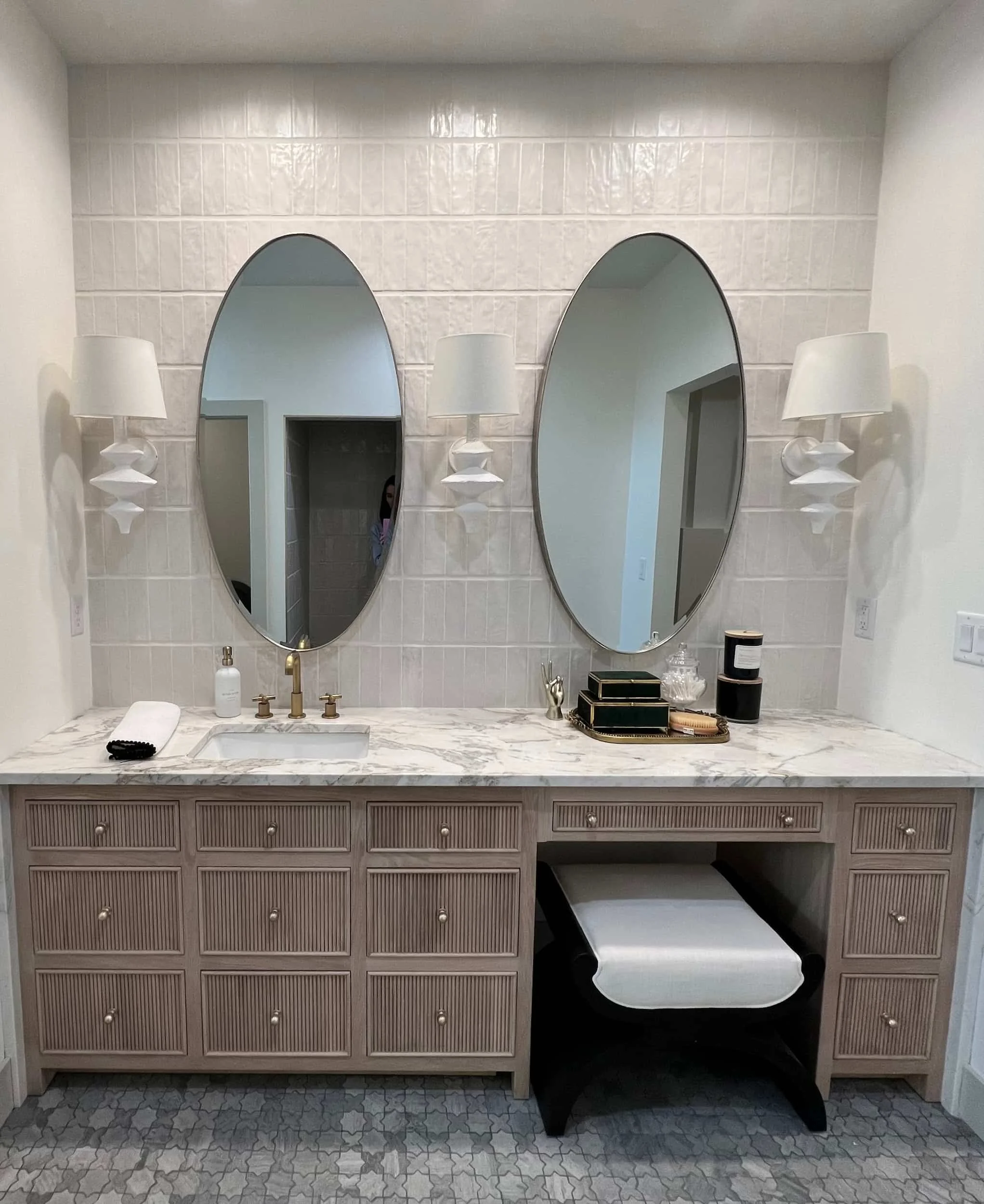 Luxurious bathroom double vanity with taupe fluted cabinetry and brass hardware. The vanity includes a central makeup area and is set against a white subway tile wall with dual oval mirrors and decorative sconces.
