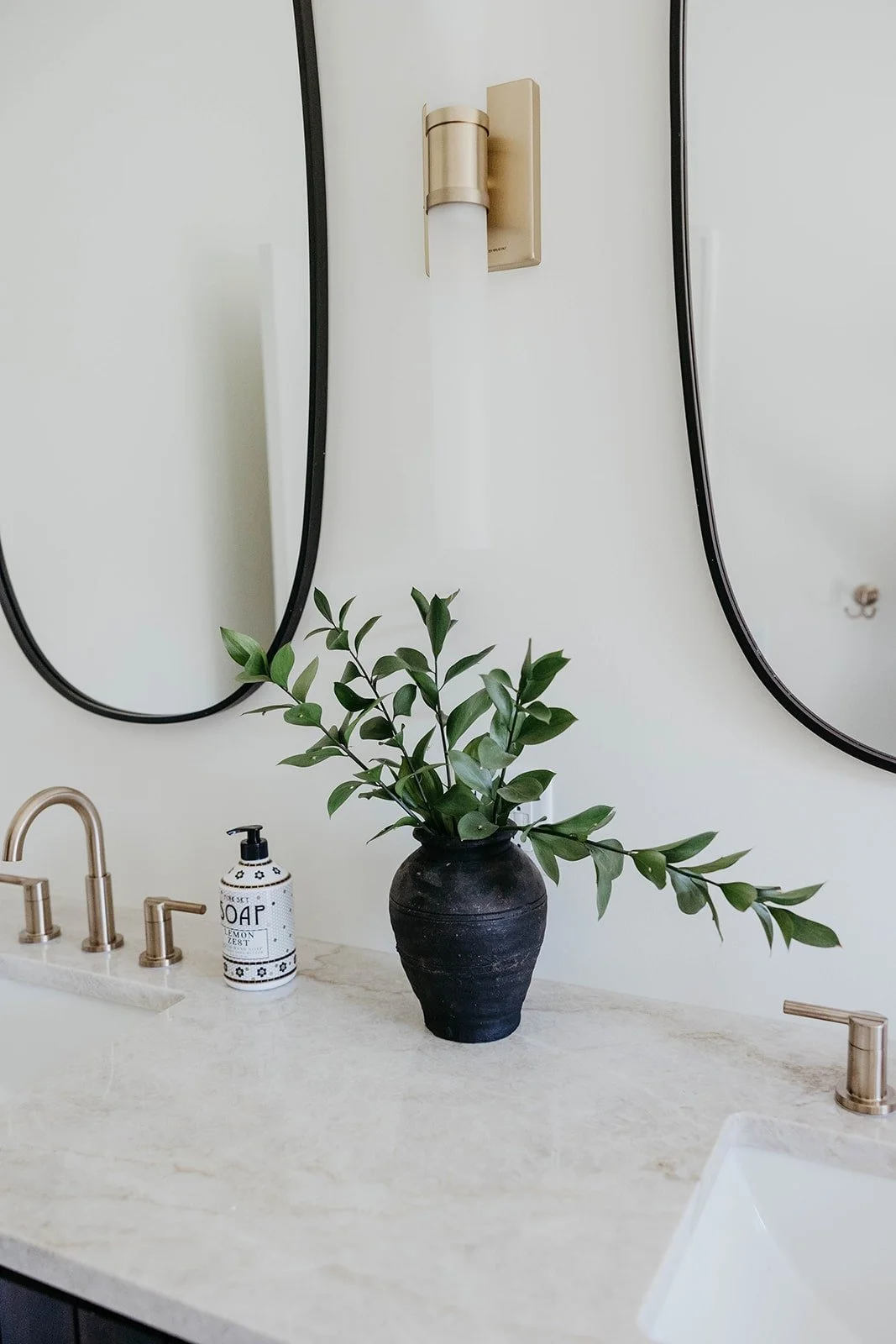Close-up of the vanity countertop styled with a black vase of greenery, a white soap dispenser, and brushed gold widespread faucets.