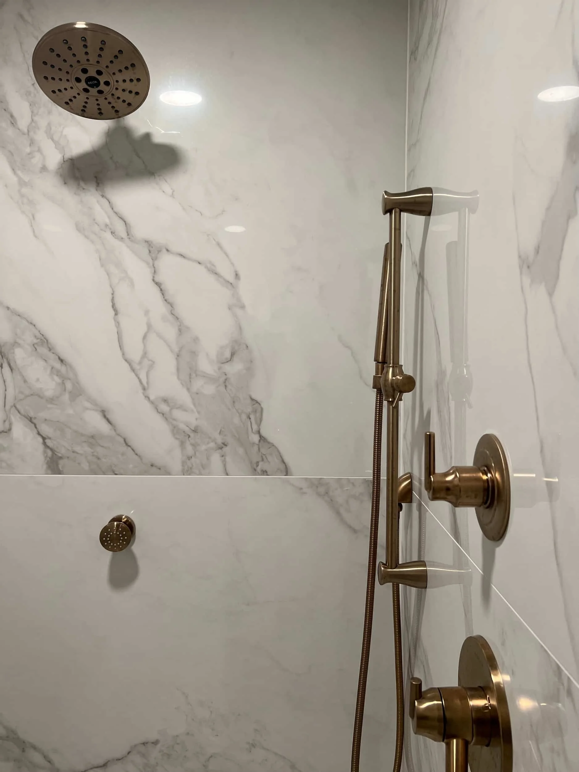 Walk-in shower interior featuring large-format white marble-look slab tile with gray veining and a complete brushed gold shower system.
