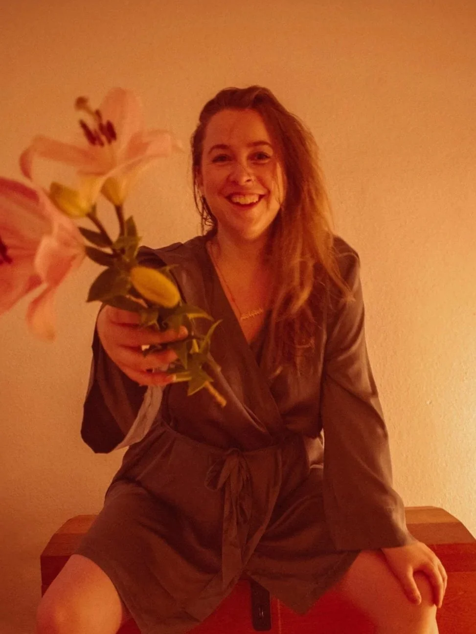 Eine lachende Frau mit langen, gewellten Haaren, die in einem dunklen Satinkimono auf einer Holzkiste sitzt, eine Lilie in die Kamera hält, vor einem warm erleuchteten, neutralen Hintergrund.