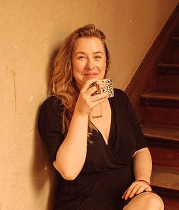 Eine lächelnde junge Frau mit lockigem, blondem Haar sitzt auf einer Holztreppe und hält eine Tasse mit Leomuster in der Hand.