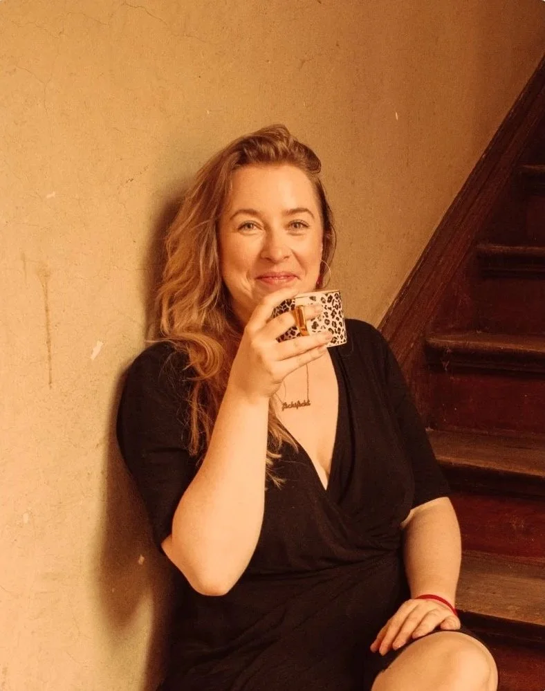 Eine lächelnde junge Frau mit lockigem, blondem Haar sitzt auf einer Holztreppe und hält eine Tasse mit Leomuster in der Hand.