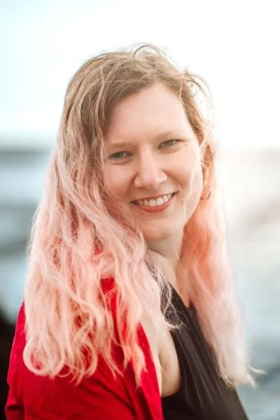 Junge Frau mit lockigem, pink-blondem Haar, lächelnd, im Freien, bei bewölktem Himmel.