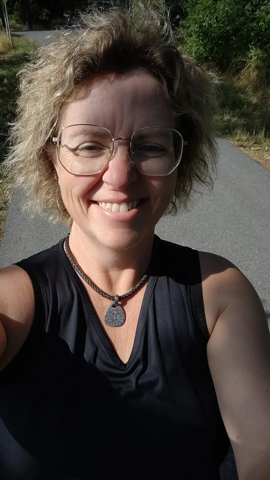 Frau macht einen Selfie im Freien bei Sonnenschein, trägt eine schwarze Sporttop und eine Halskette mit Anhänger, hat lockiges blondes Haar und trägt große Brillengläser.