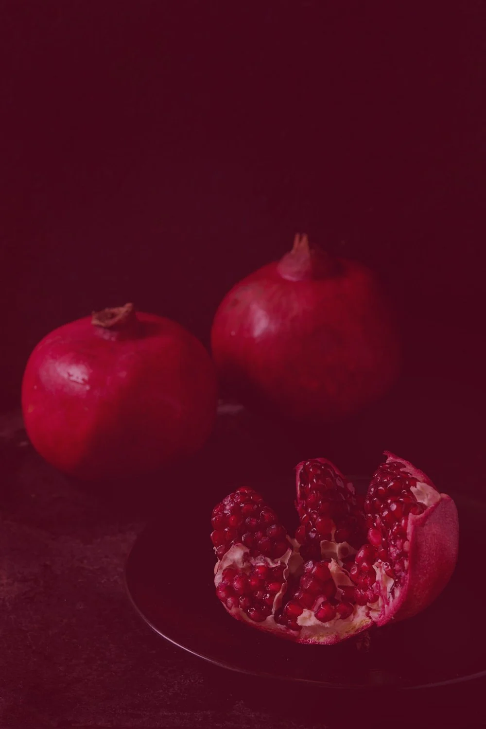 Drei Granatäpfel, wobei einer halb geöffnet ist und auf einem schwarzen Teller liegt, dunkler Hintergrund, rotes Licht.