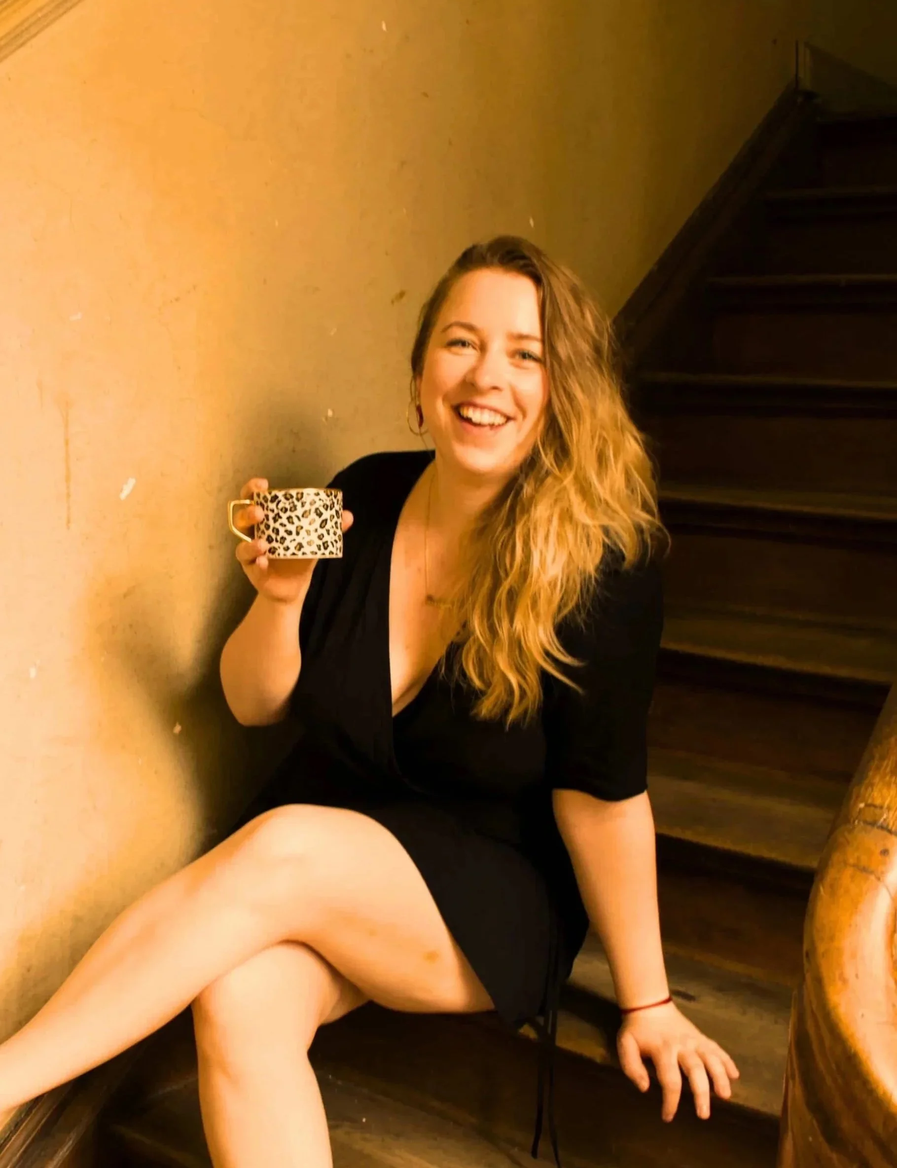 Lächelnde junge Frau mit langem braunen Haar sitzt auf einer Holztreppe, hält eine Leoparden-Print-Tasse, trägt ein schwarzes Kleid und hat ein rotes Armband am Handgelenk.