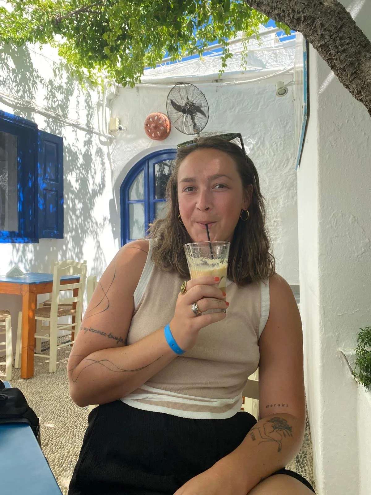 Eine junge Frau mit braunen, lockigen Haaren trinkt einen cremigen Drink mit einem Strohhalm in einem Café mit weißer Wand und blauen Fenstern, im Freien bei Sonnenlicht, umgeben von grünen Bäumen.