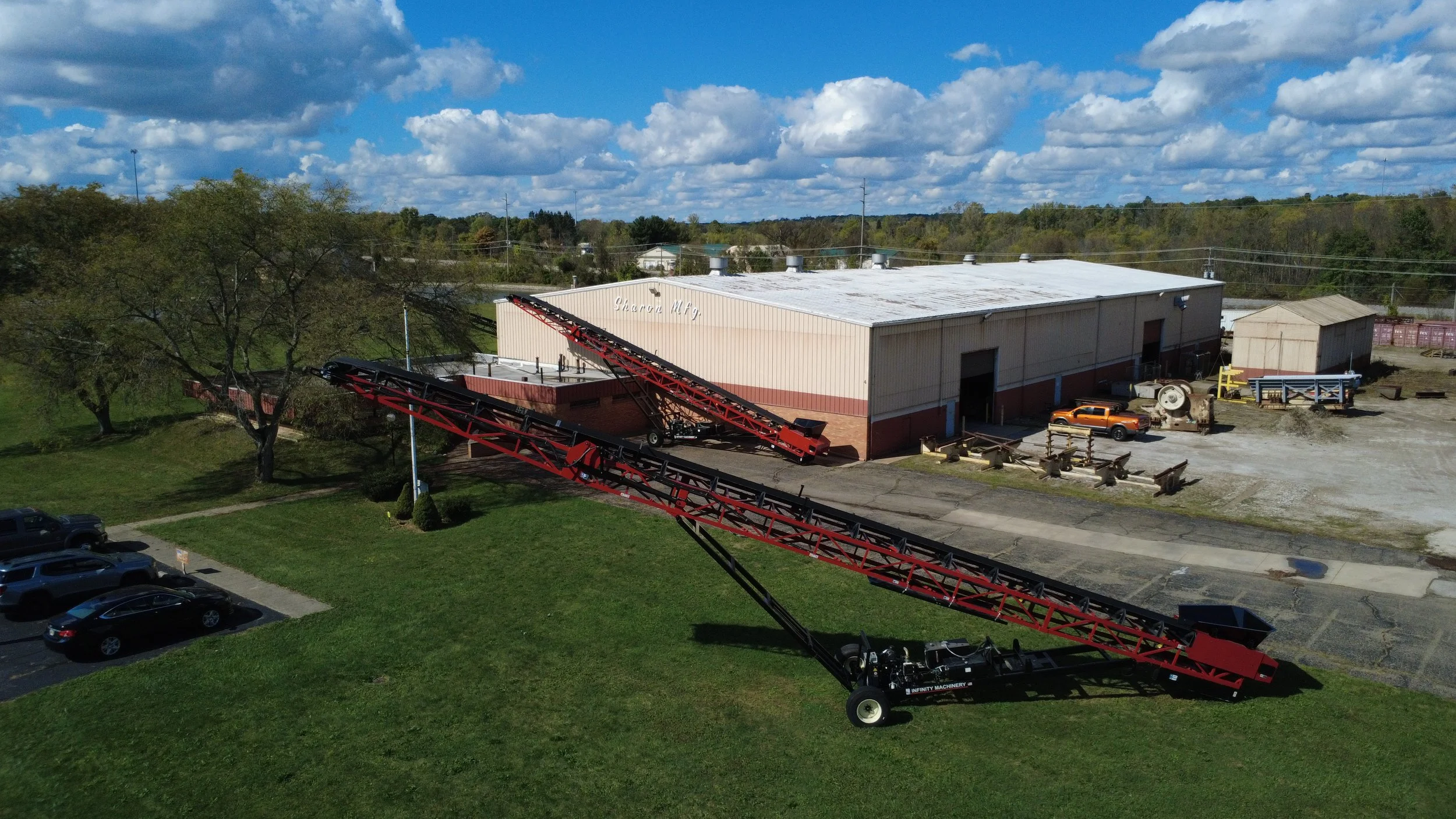 An aerial view of an industrial area with a warehouse, machinery, and vehicles, including a large red and black conveyor system on a grassy area.