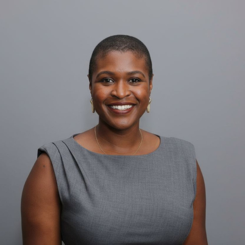 Portrait of a smiling Black woman with a short buzz cut, wearing a gray sleeveless dress and gold earrings against a gray background.