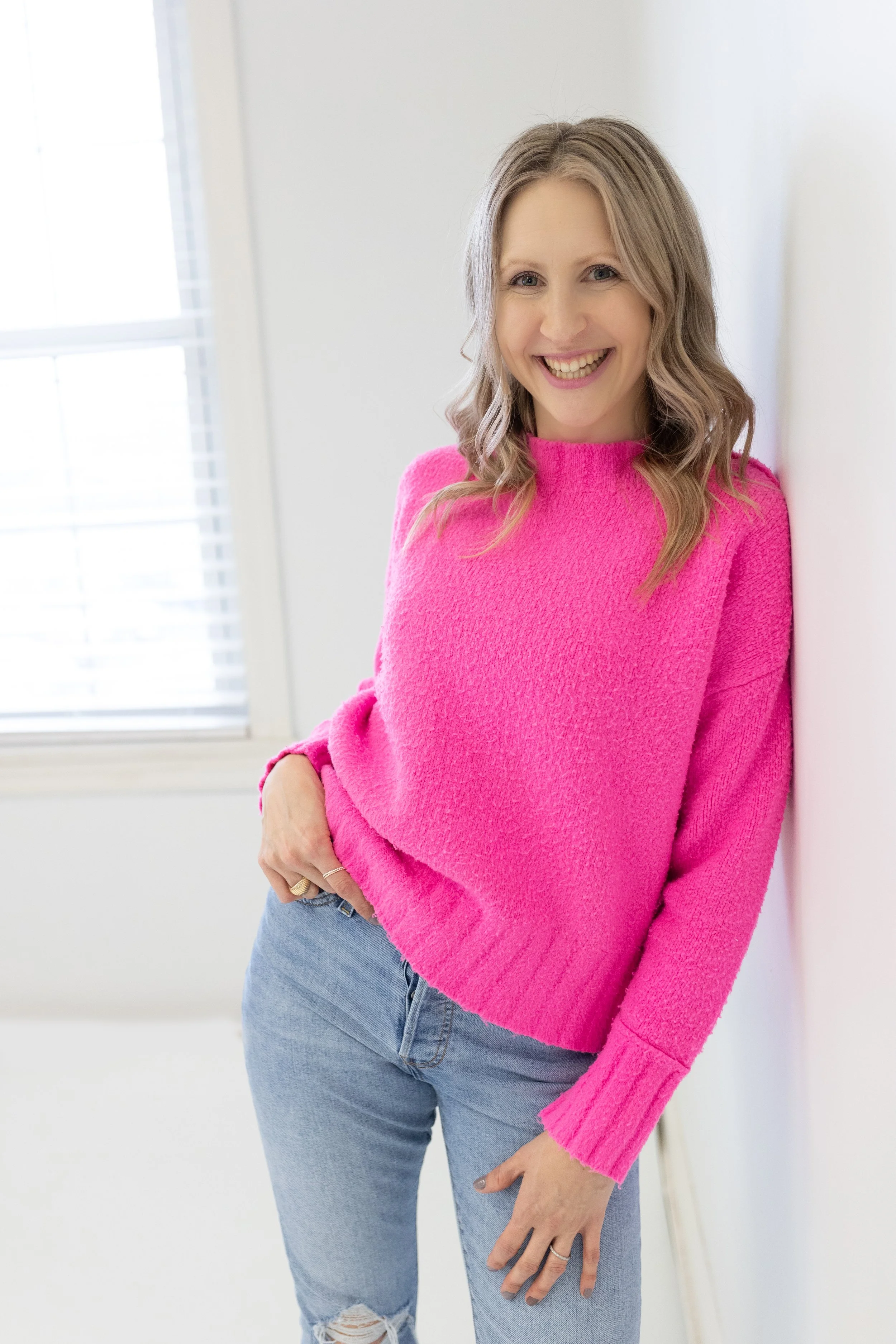 A woman with blonde wavy hair smiling while leaning against a white wall in a brightly lit room, wearing a pink sweater and light blue jeans.