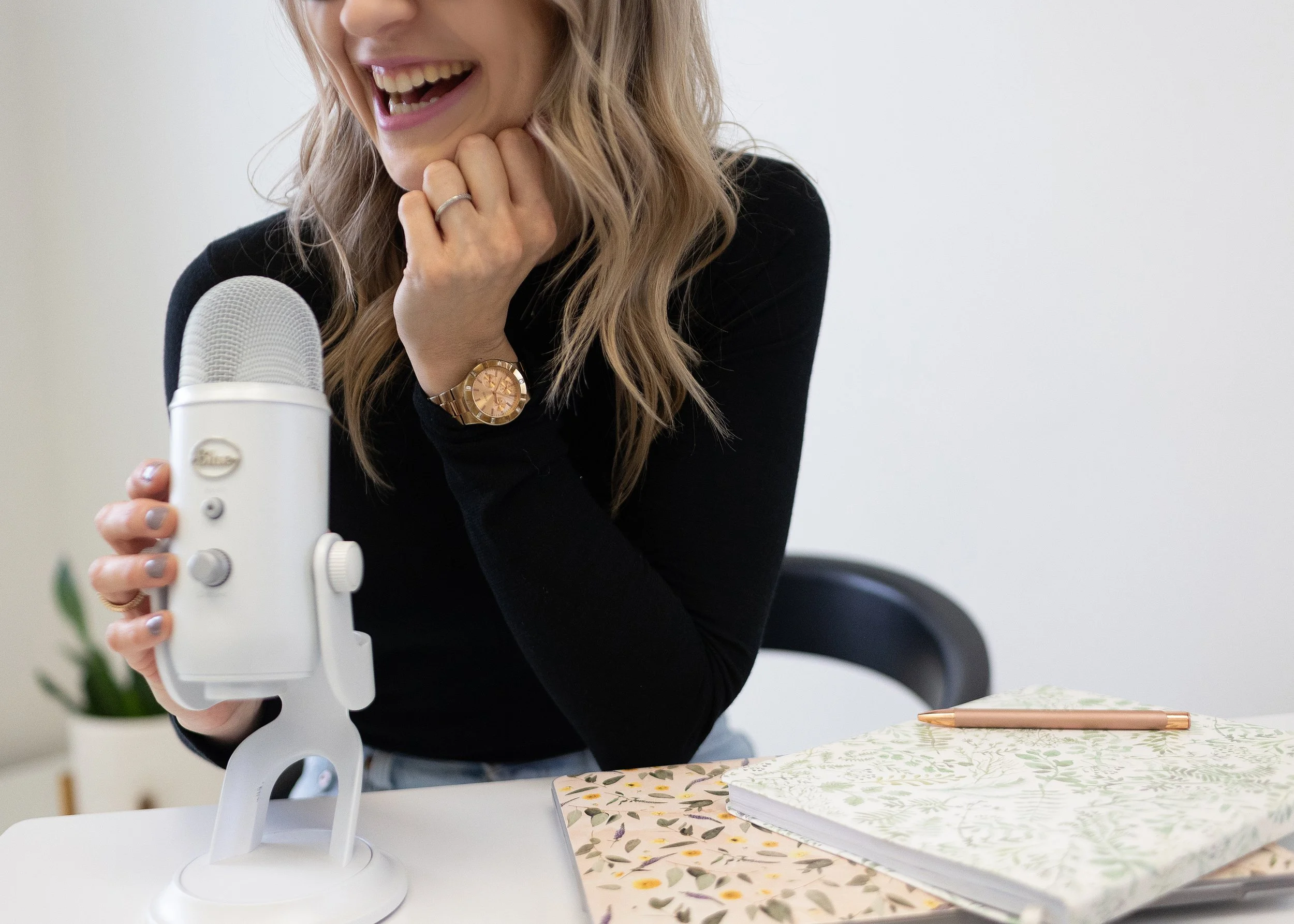 A woman with blonde hair, smiling and laughing while holding a white microphone on a table. She is wearing a black top, a gold watch, and a ring. The table has notebooks and a pen, with a plant in the background.
