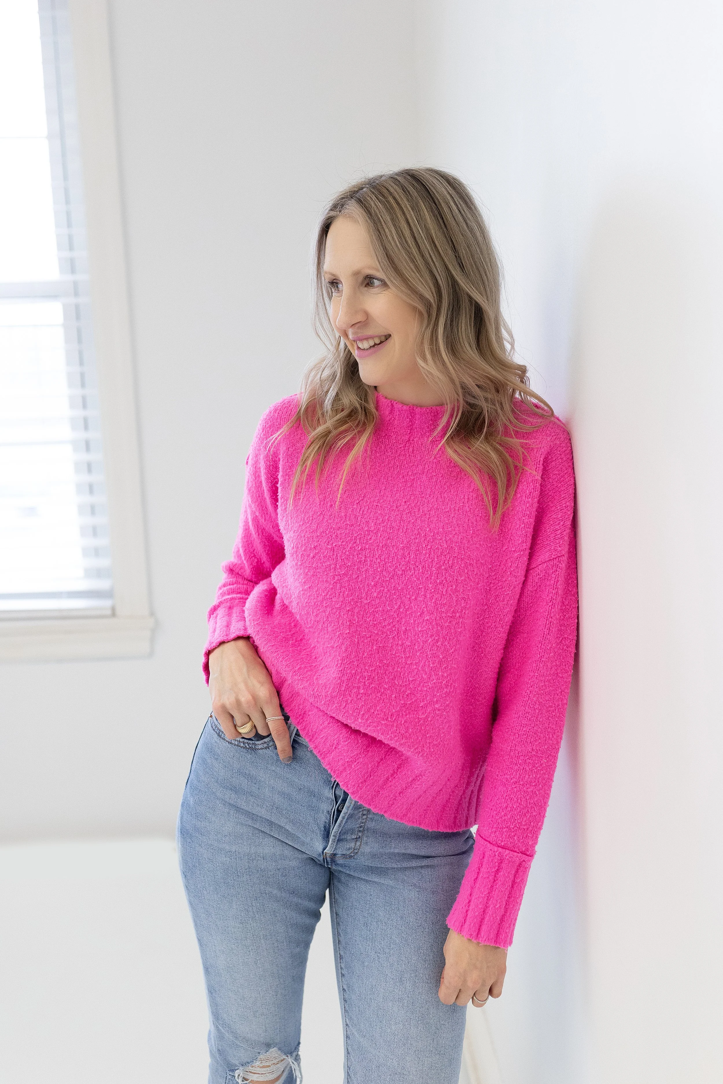 A woman with light brown, wavy hair wearing a bright pink sweater and ripped jeans, smiling and standing indoors near a white wall, with sunlight coming through a window.