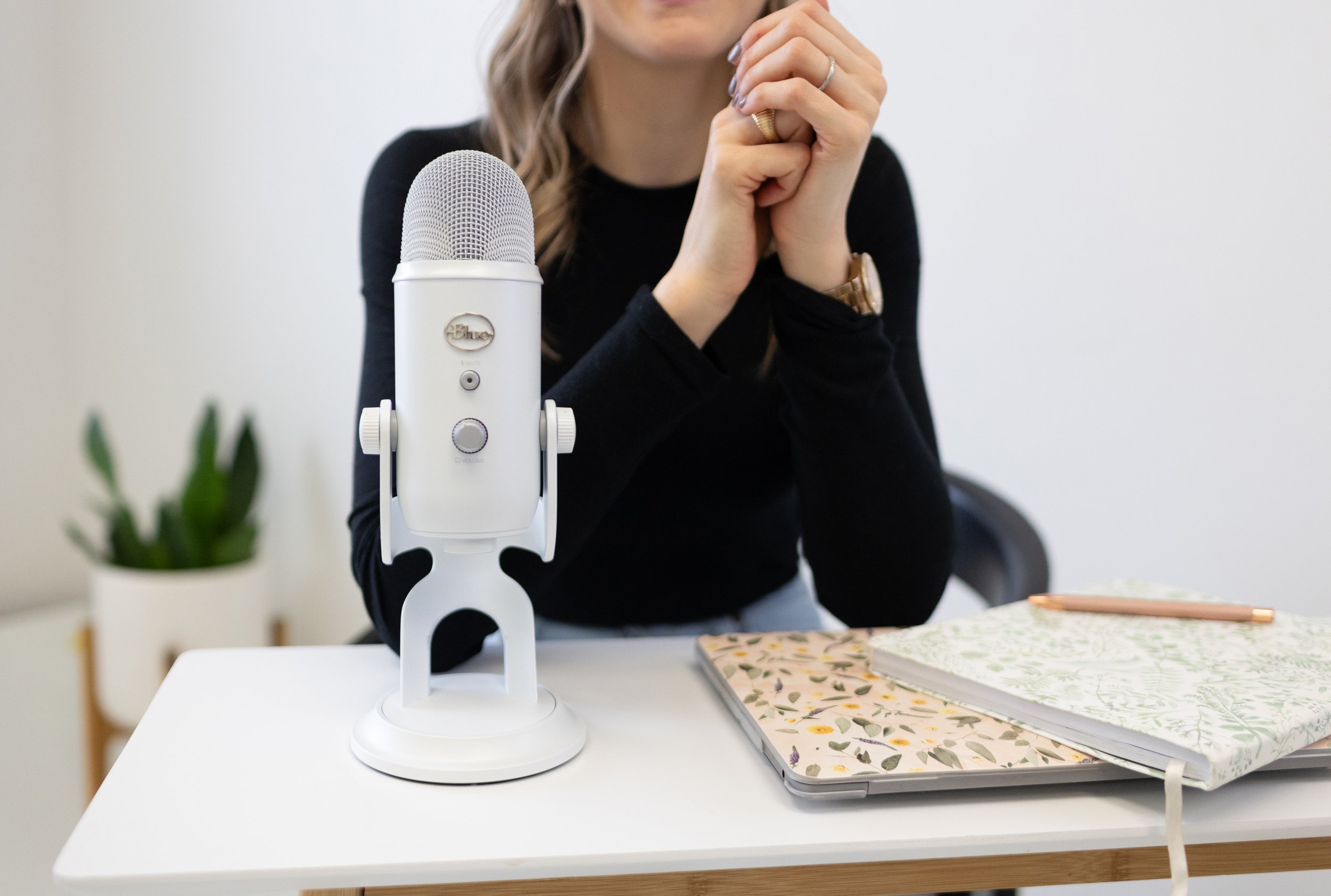 A woman sitting at a white desk with a white microphone, a laptop, notebooks with floral covers, and a pen. She has her hands clasped near her face and is wearing a black top, a watch, and rings. In the background, there's a potted plant.