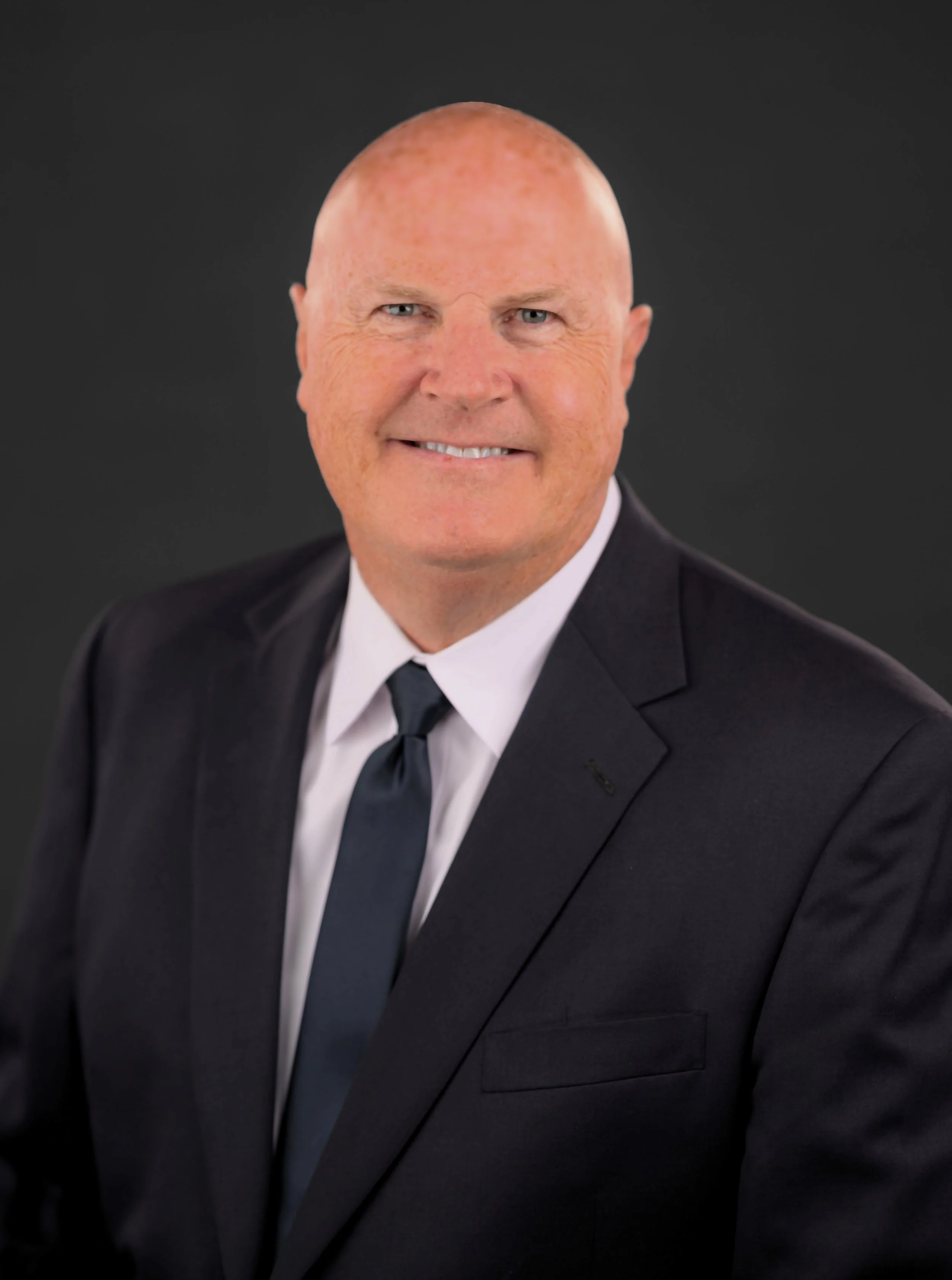 A professional engineer in a dark suit, white shirt, and dark tie, smiling against a dark background who teaches flight control design.