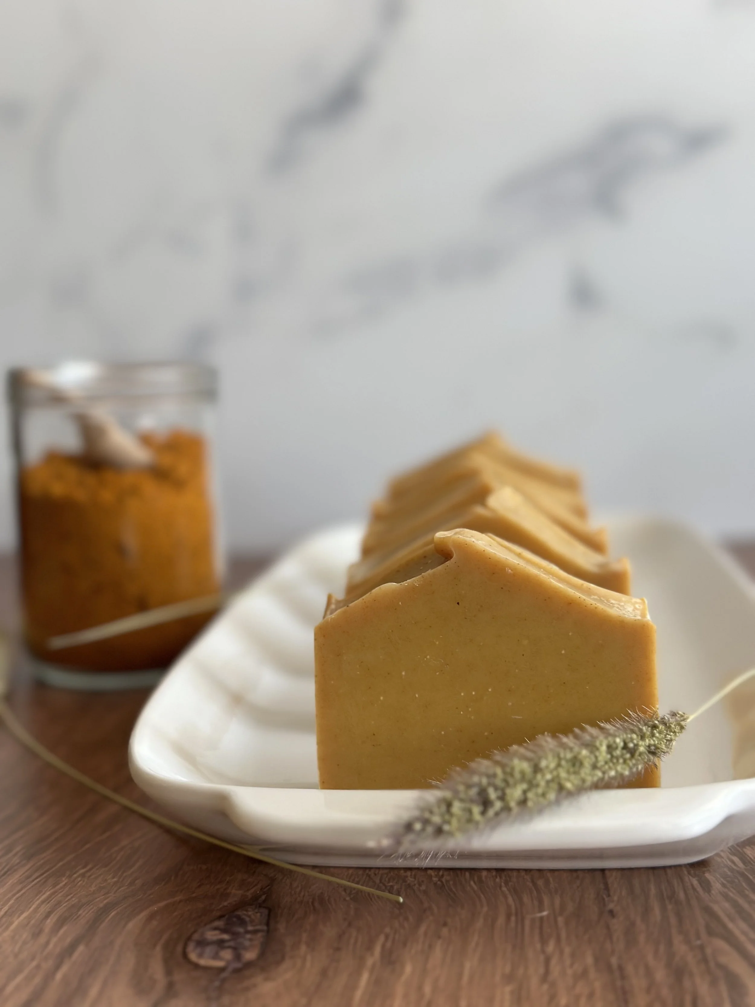 A white porcelain dish with slices of caramel-colored soap, placed on a wooden surface, with a decorative dry plant and a jar of brown substance in the background.