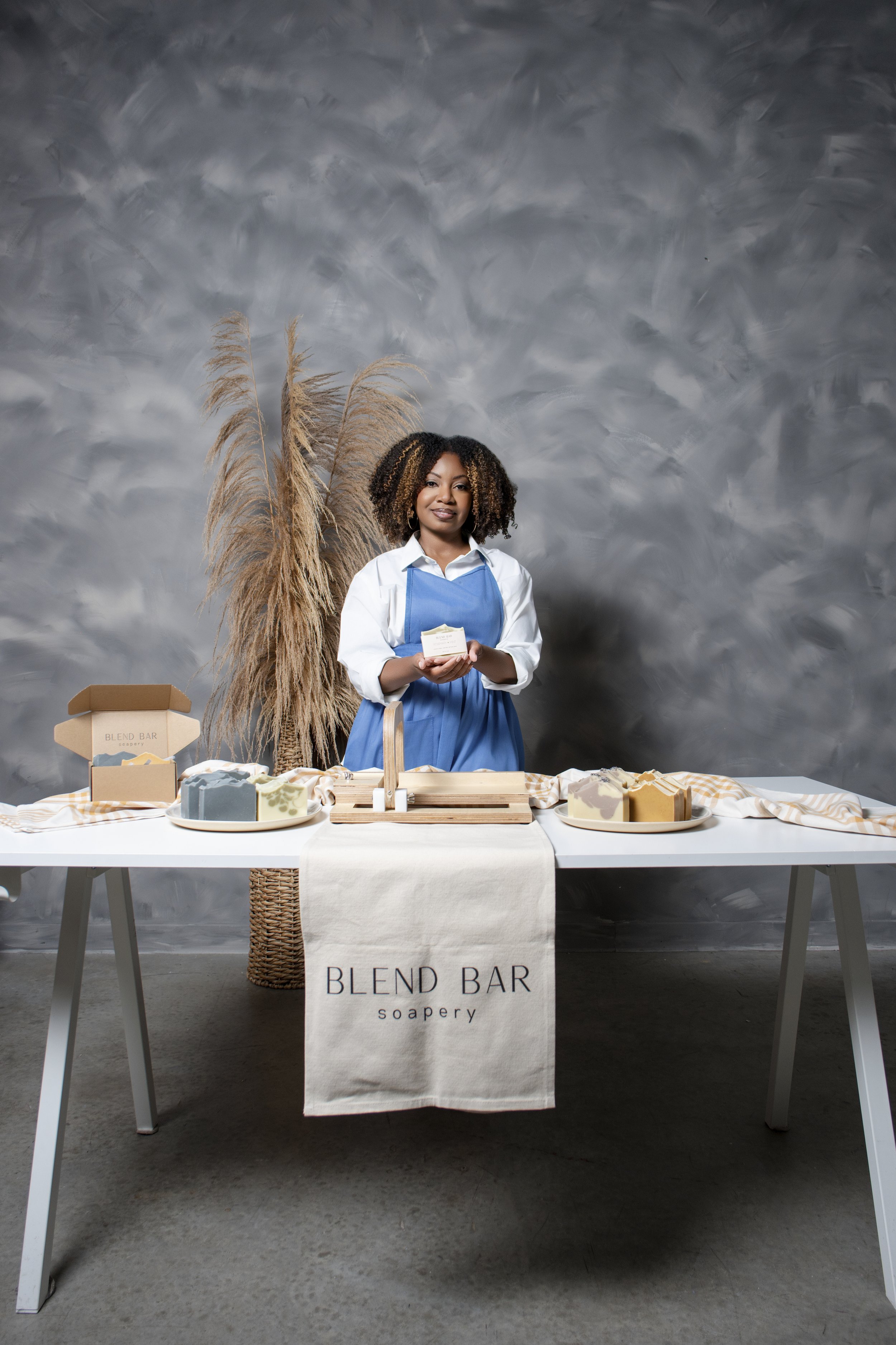 A woman in a blue apron holding soap in front of a table with soap displays at a soapery called 'Blend Bar'. The table has a white cloth with the soapery's name on it, and there are various soap bars and packaging on the table. The background features a decorative potted pampas grass and a textured gray wall.