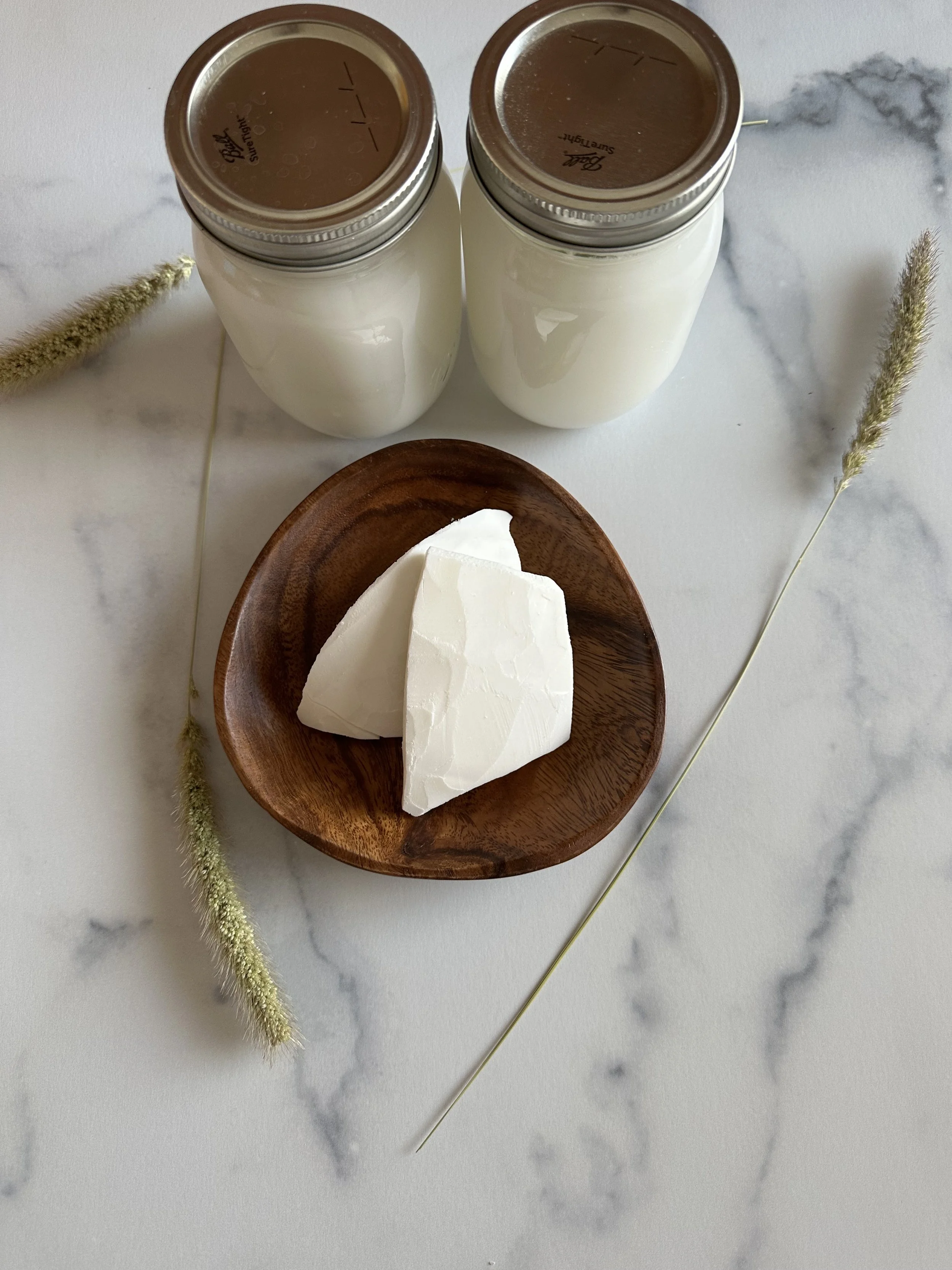 Two glass jars of milk with metal lids, a wooden bowl with white blocks of cheese, and decorative dried grass on a marble surface.