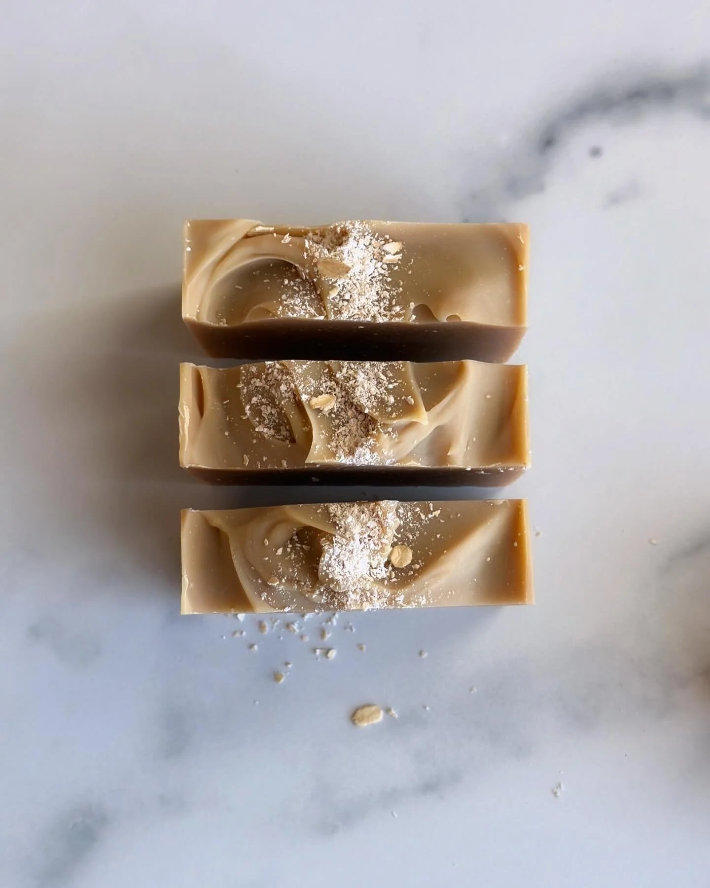 Three pieces of beige and brown swirled soap with white powder sprinkled on top, placed on a white marble surface.