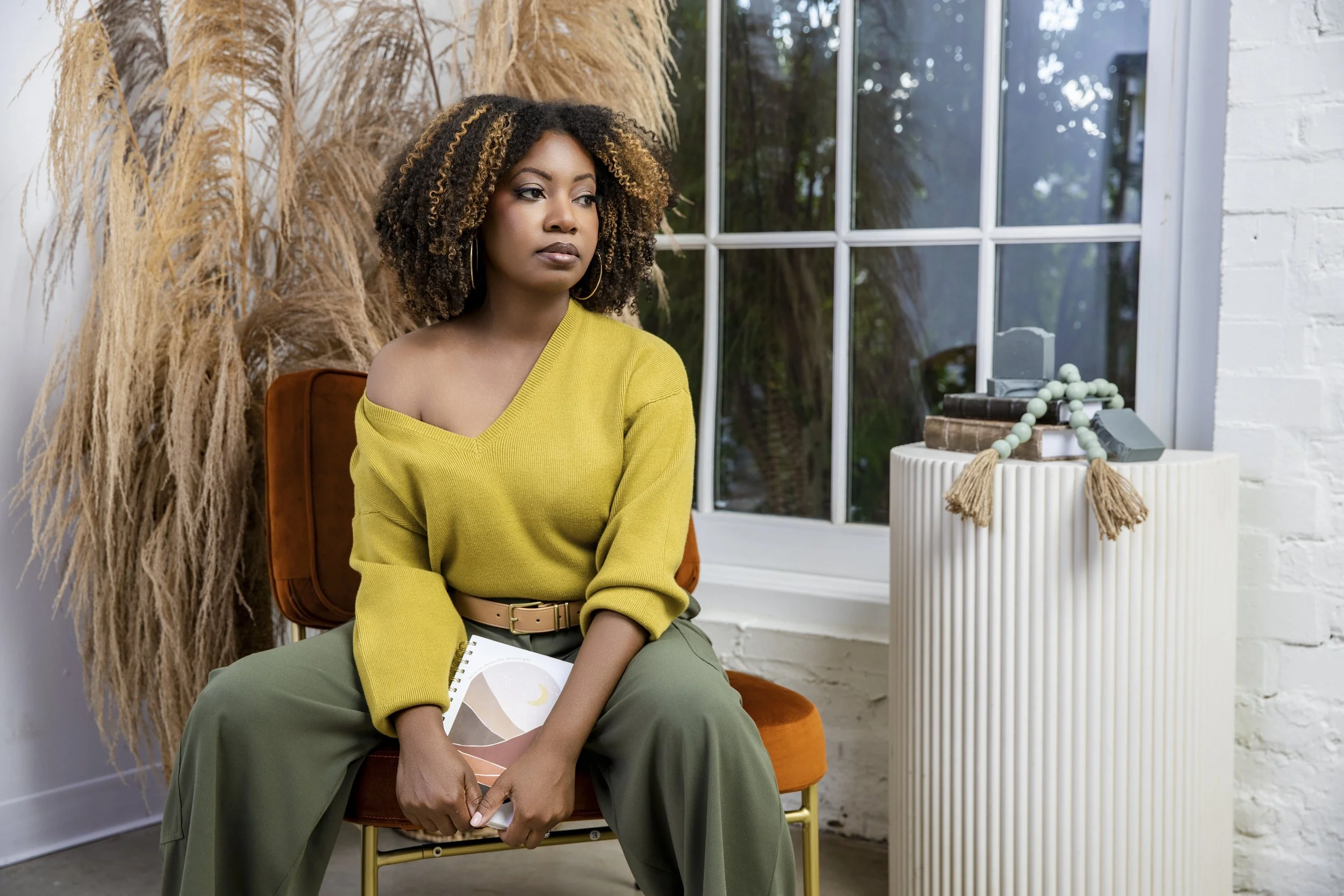 A woman with curly hair wearing a yellow off-shoulder sweater and green pants sitting on an orange chair holding a notebook, inside a room with large window and decorative items on a white pedestal.