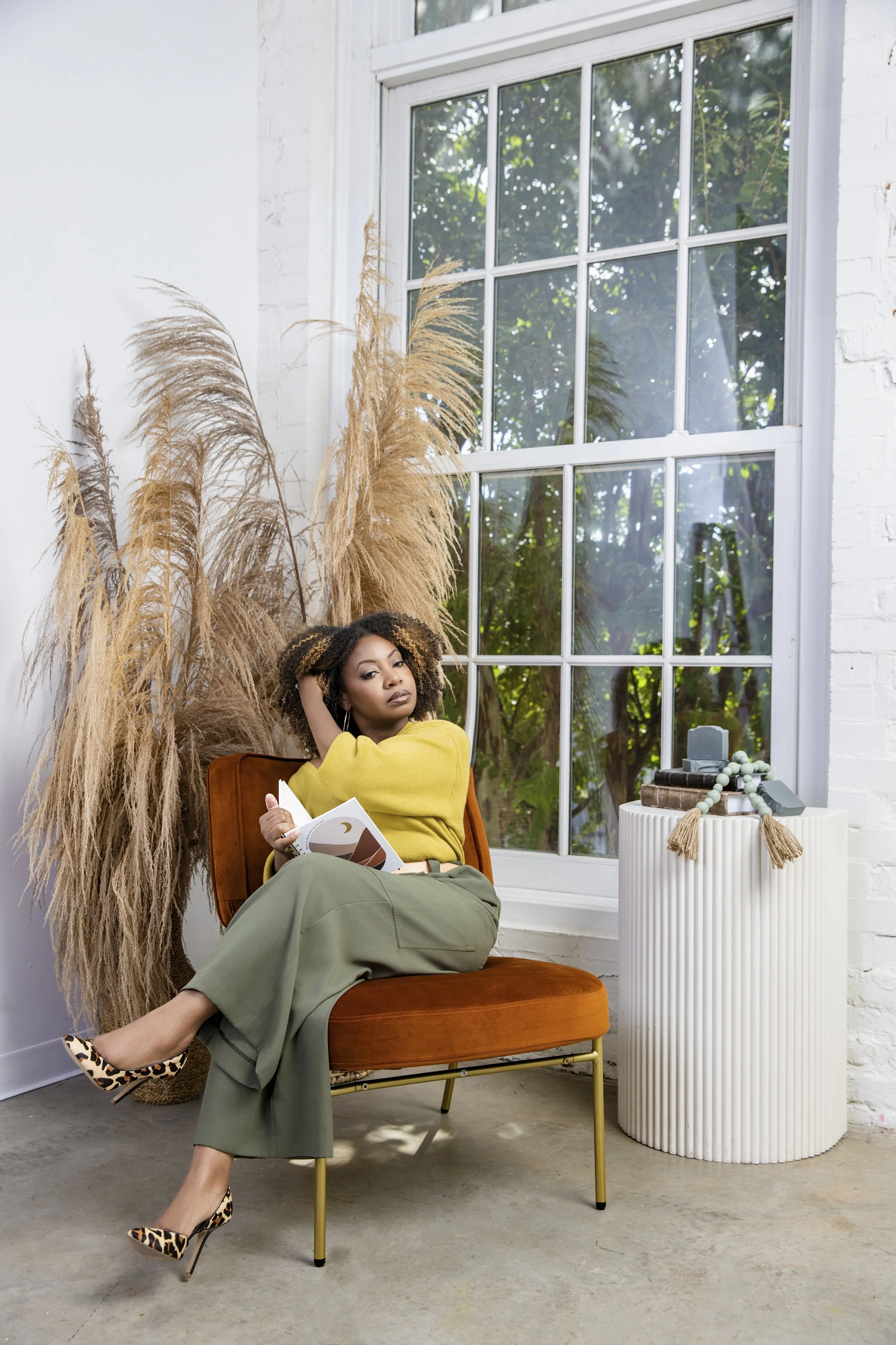 A woman with curly hair sitting in a modern room, reading a book, wearing a yellow sweater, gray wide-leg pants, and leopard print heels, with dried pampas grass behind her and a white side table with decorative items beside her.