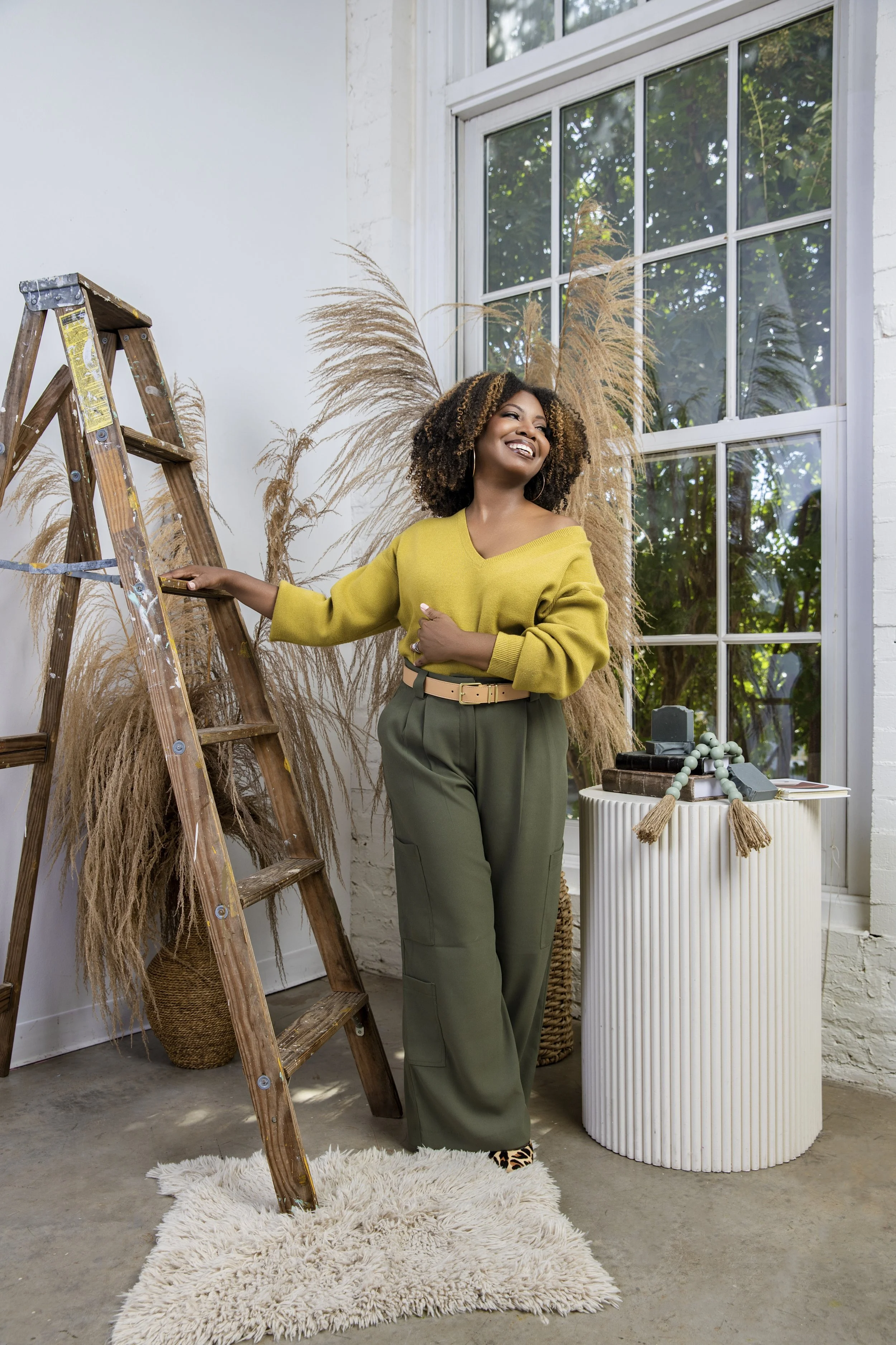 A woman with curly hair, smiling and wearing a yellow sweater and green pants, standing beside a wooden ladder in a room with large windows and decorative brown plants.