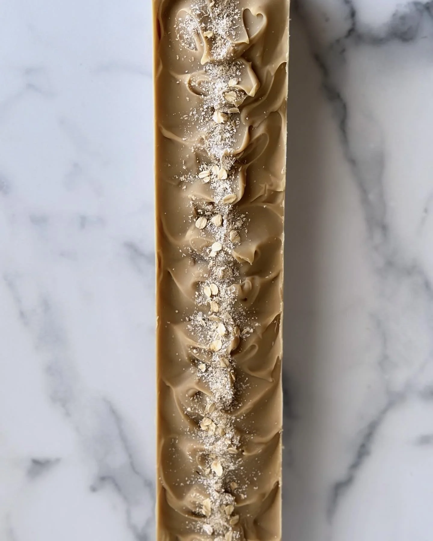 Top view of a long, narrow caramel chocolate bar decorated with oats, powdered sugar, and swirl patterns on a white marble surface.