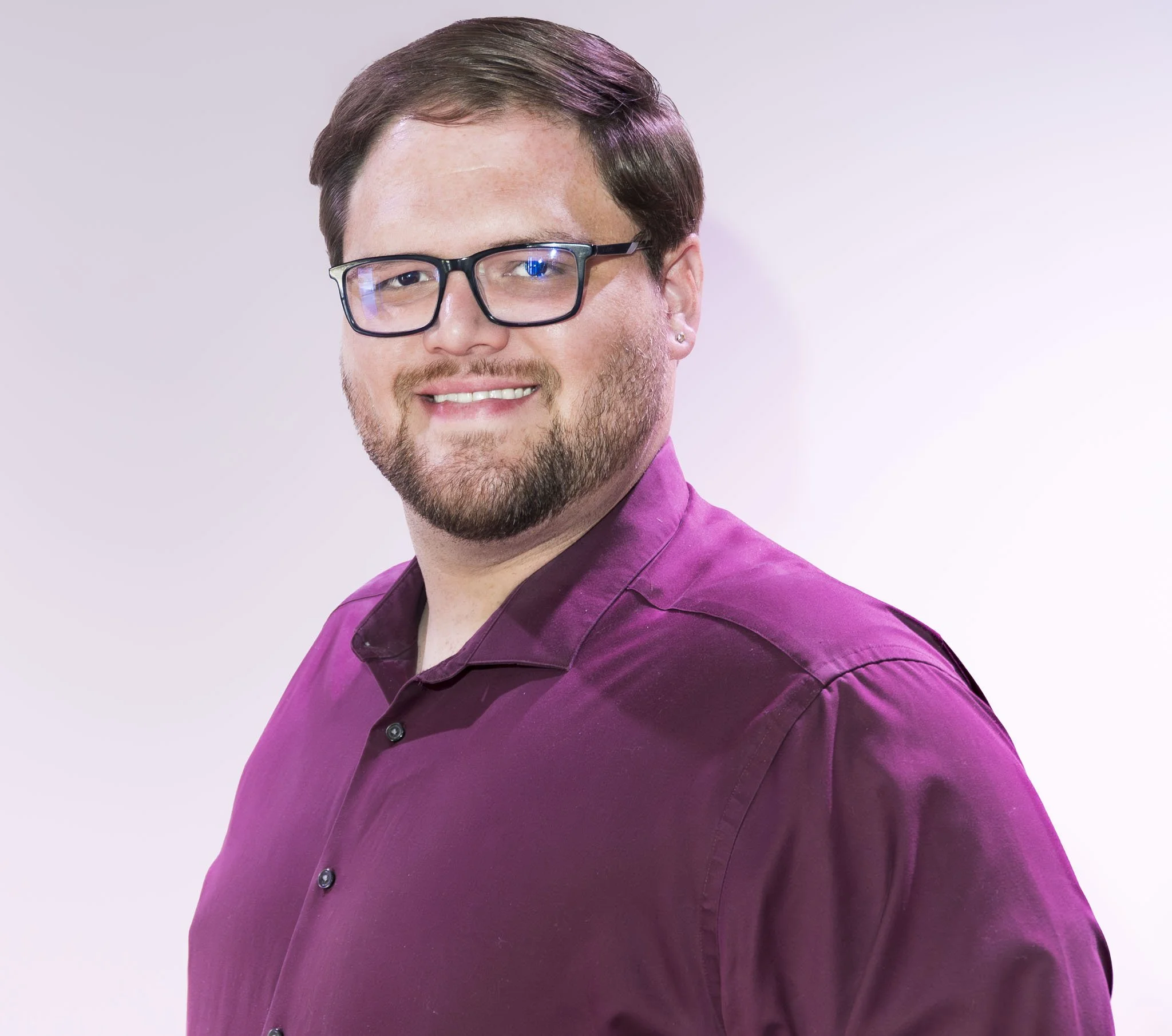 Portrait of a smiling man with glasses, a beard, wearing a purple shirt, against a plain light background.