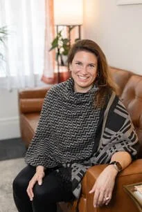 A picture of a light skinned woman with long brown hair, smiling, with a gray patterned shawl across her shoulders