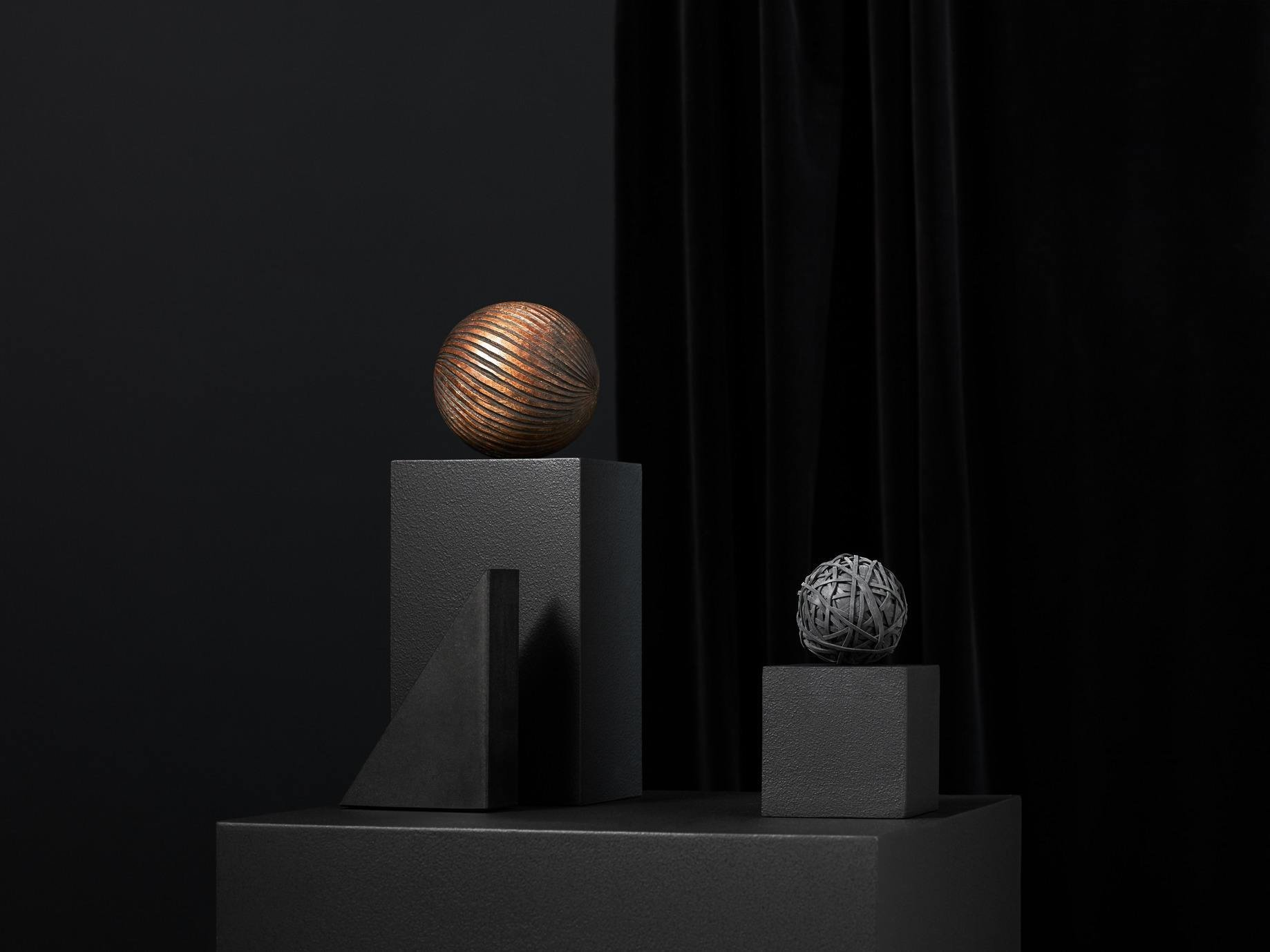 Two geometric sculptures, one with a striped sphere on a rectangular pedestal, the other with a tangled spherical object on a smaller cube, set against a dark background with black curtains.