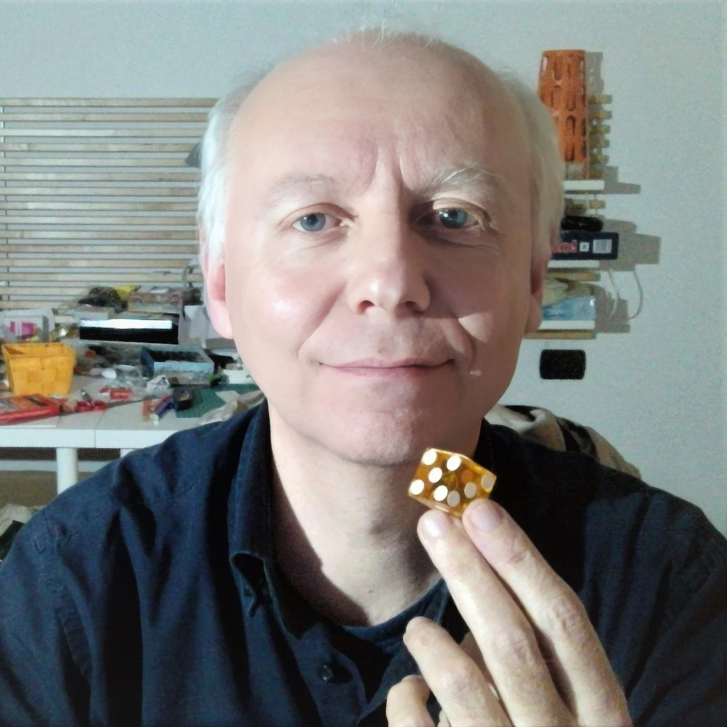 Uomo con capelli biondi e occhi azzurri tiene una miniatura di dado dorato, sfondo di una stanza con mensole e strumenti di lavoro.