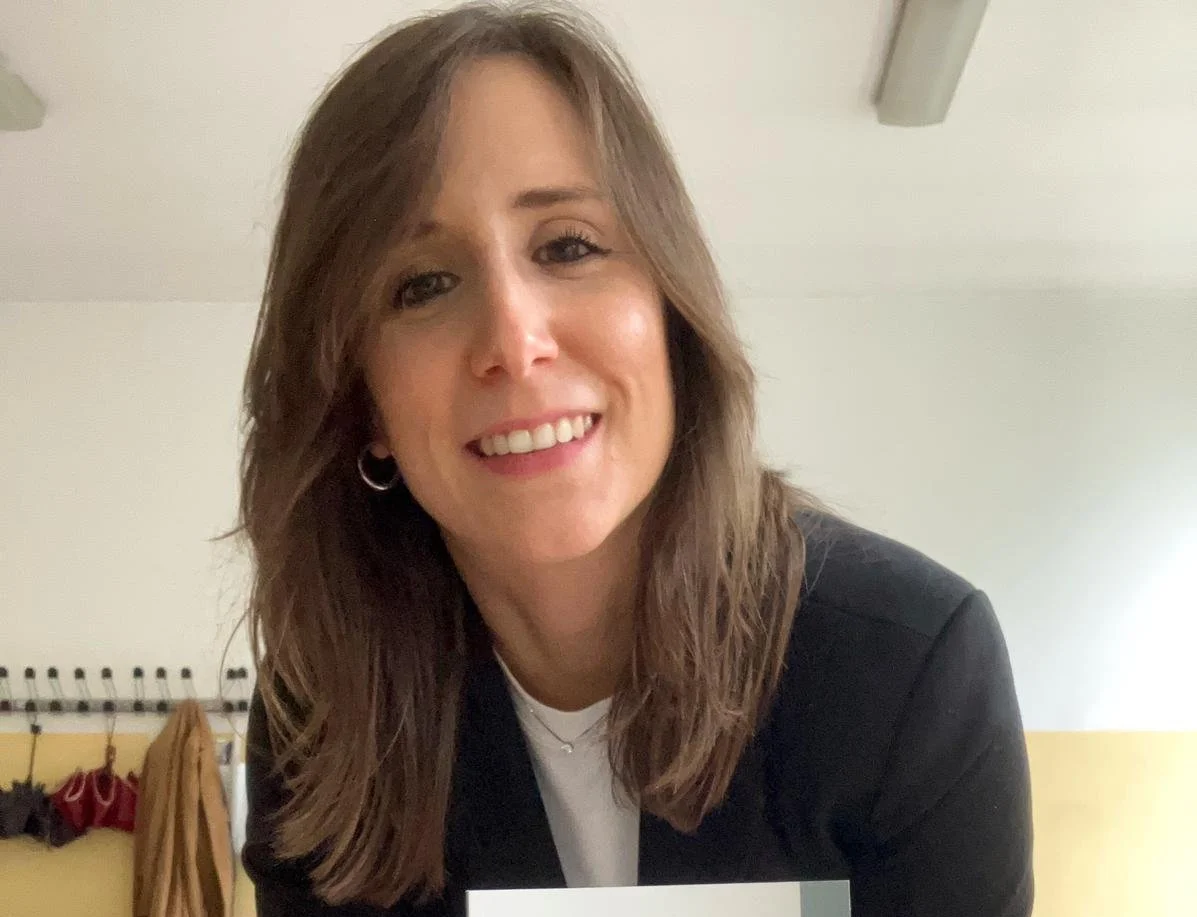 Una donna sorridente con capelli castani, indossa una maglietta bianca e una giacca nera, in un ambiente interno con pareti chiare.