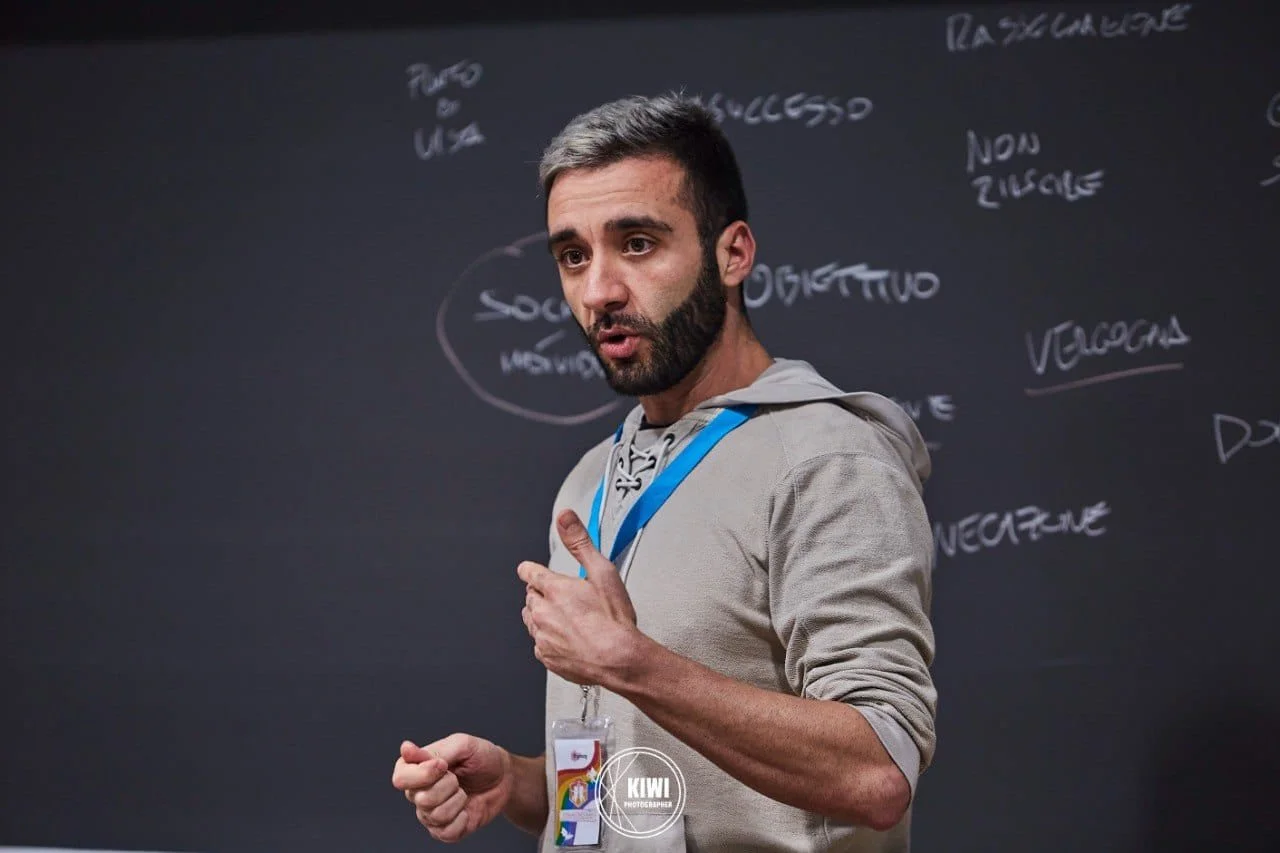 Un insegnante davanti a una lavagna con scritte in italiano, che spiega concetti a una lezione, con un badge al collo e un cappotto grigio.