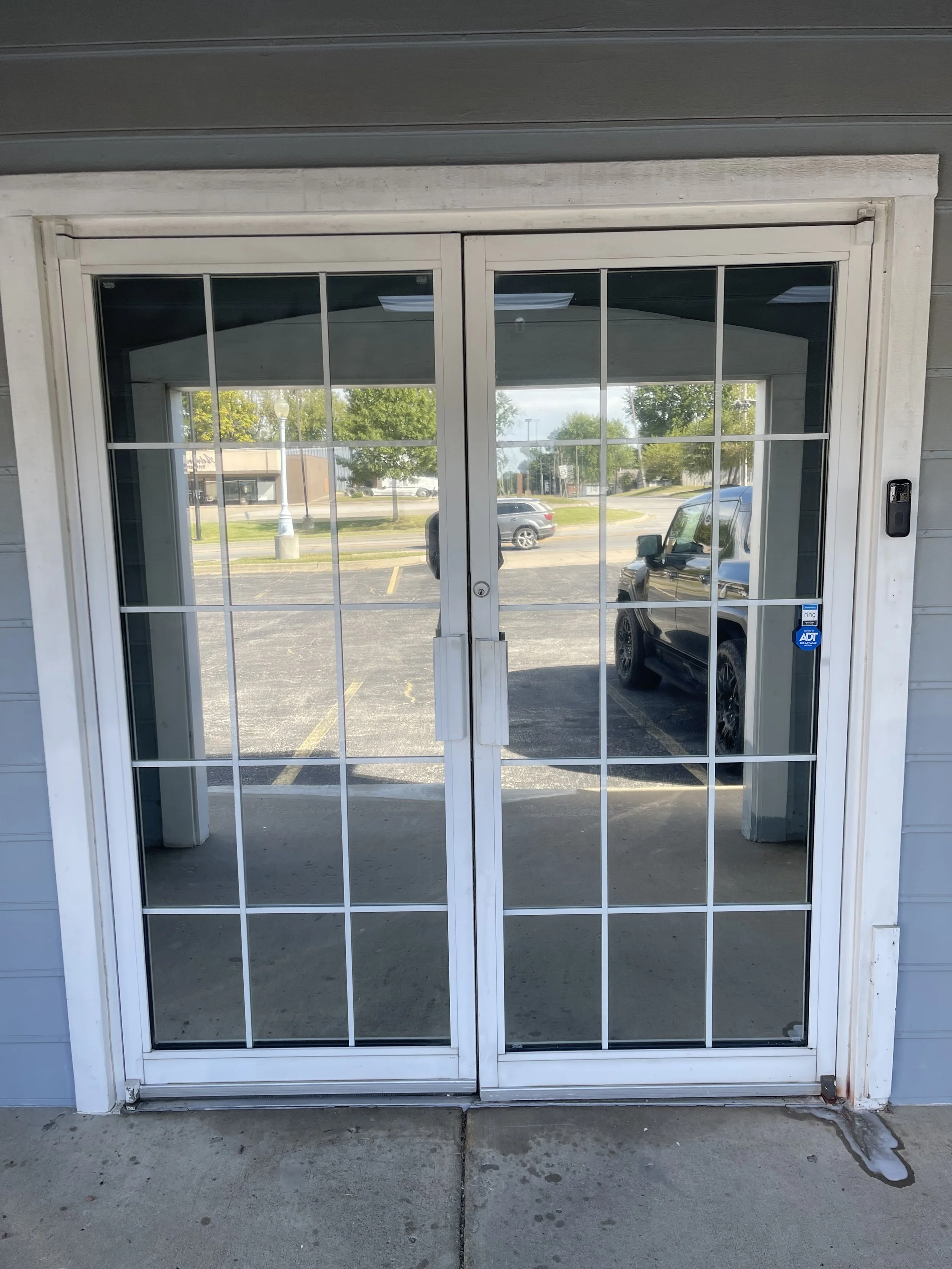 Glass double door with white grid pattern, framed in white, with an electronic access keypad on the right side, glass panels reflecting a parking lot and trees outside.