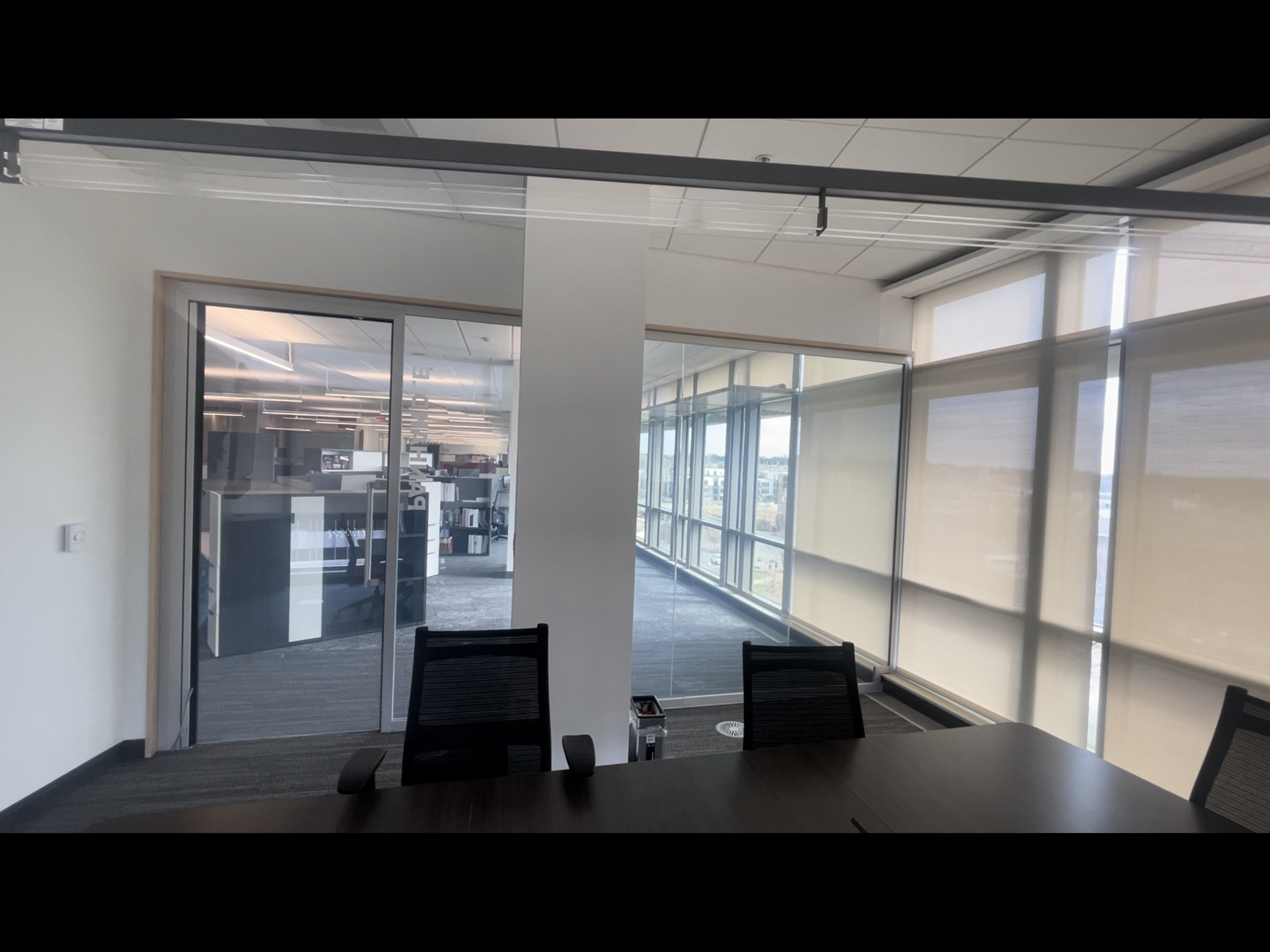 Office conference room with glass walls, office chairs, and large windows with shades, view of city buildings outside. Smart Film powered on. 