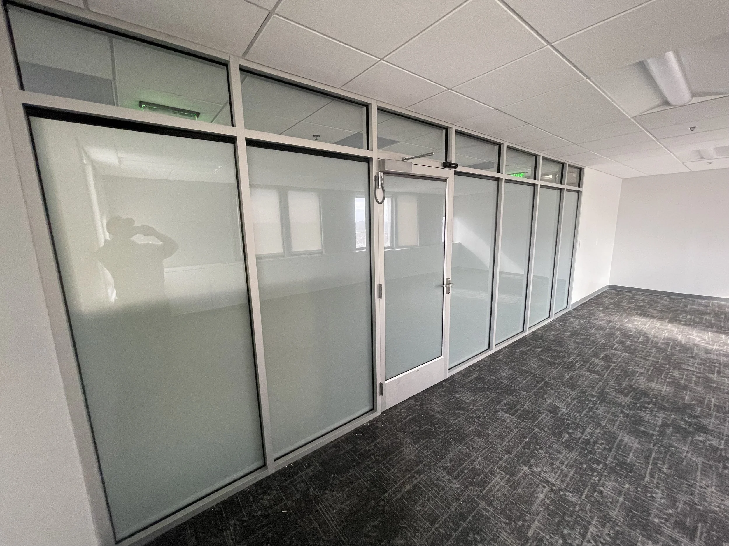 Empty office space with frosted glass walls and a door, fluorescent ceiling lights, and a dark patterned carpet.