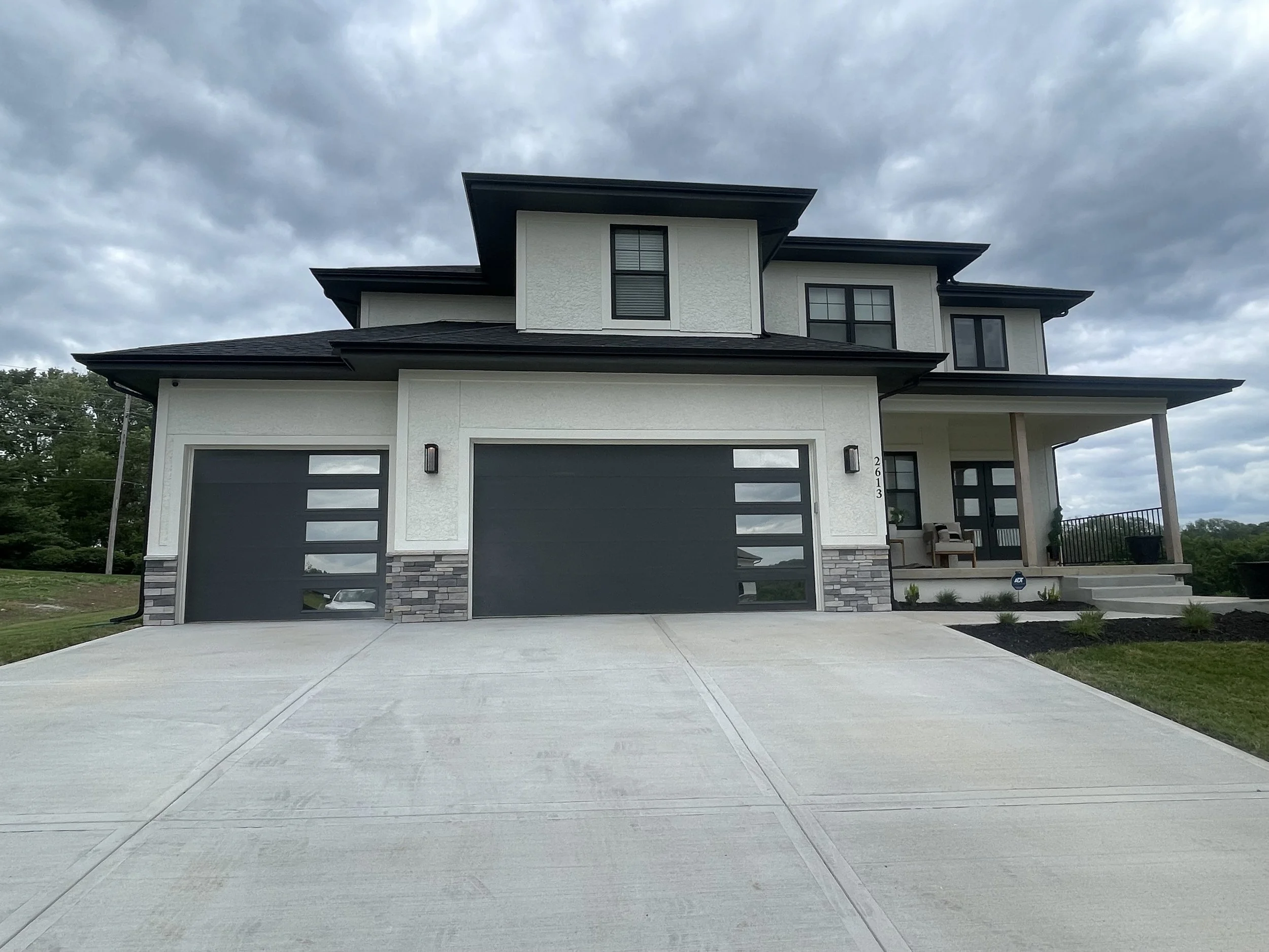 Modern two-story house with a white exterior and black roof, featuring two large garage doors with rectangular windows, a porch with seating, and a driveway leading up to it, under a cloudy sky.