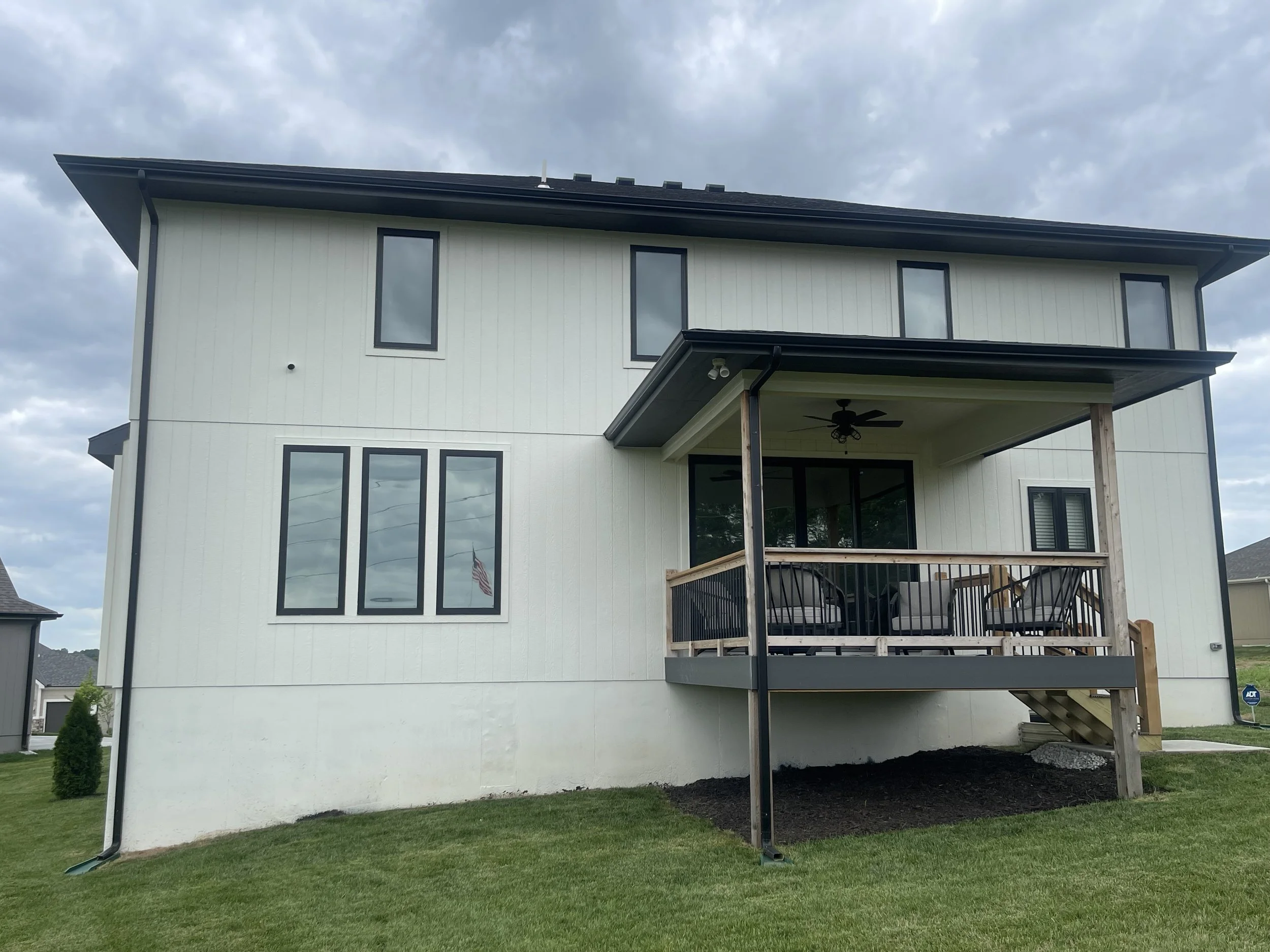 Back view of a two-story white house with a covered balcony and a ceiling fan, surrounded by a green lawn with a small shrub, under a cloudy sky.