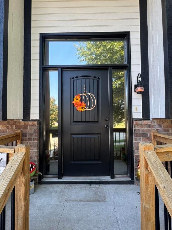 Front door of a house with a black door decorated with a pumpkin-shaped wreath with orange flowers, flanked by side windows. A porch with wooden railing and steps, and a brick and siding exterior wall.