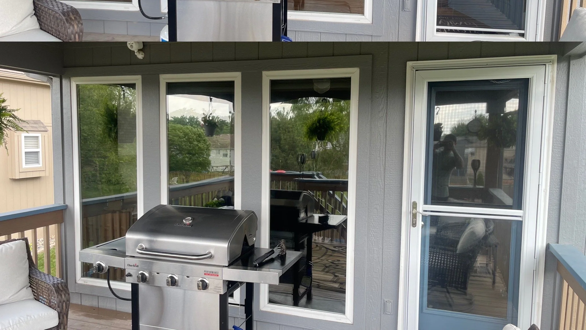 A stainless steel propane grill on a porch with a sliding glass door and windows; balcony railing, cushioned wicker chair, and reflections of a person in the glass.