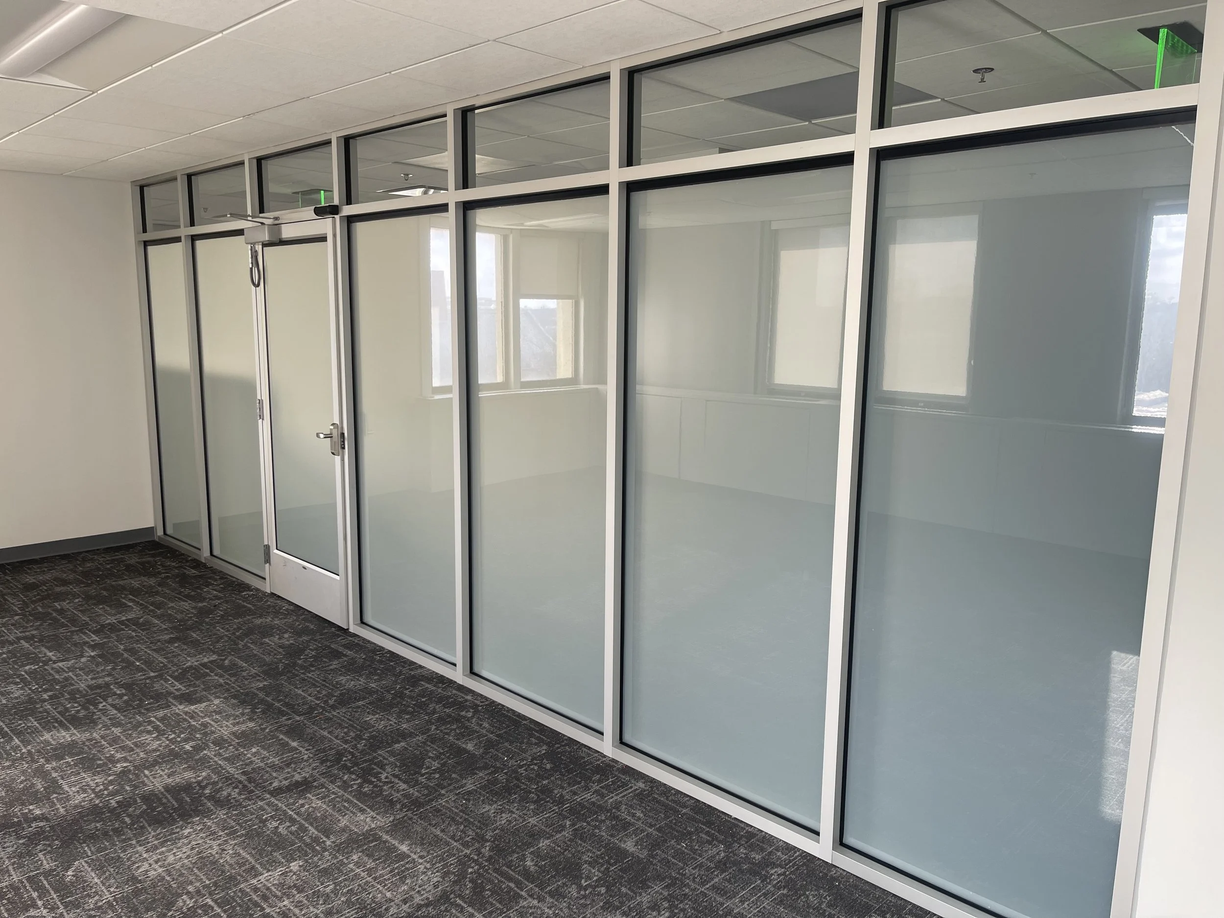 Office interior with glass partition walls and door, carpeted floor, and windows visible through the glass.