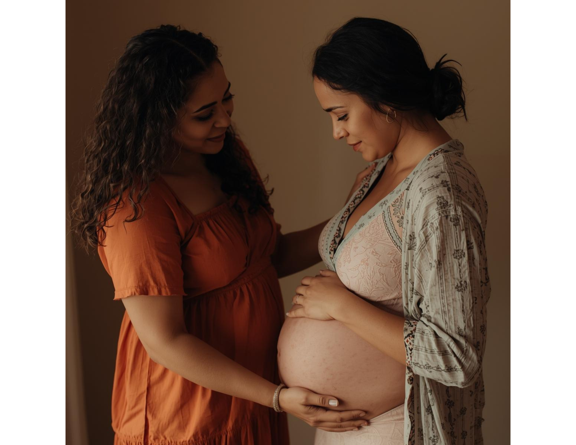 Pregnant woman with dark hair in a bun holding her belly, smiling softly, with a woman with curly hair wearing an orange dress touching her belly.