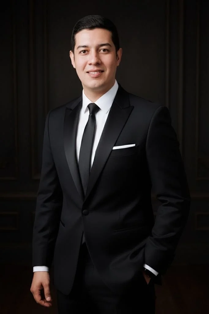 Joven vestido con traje negro, camisa blanca y corbata negra, posando con una sonrisa en un fondo oscuro.