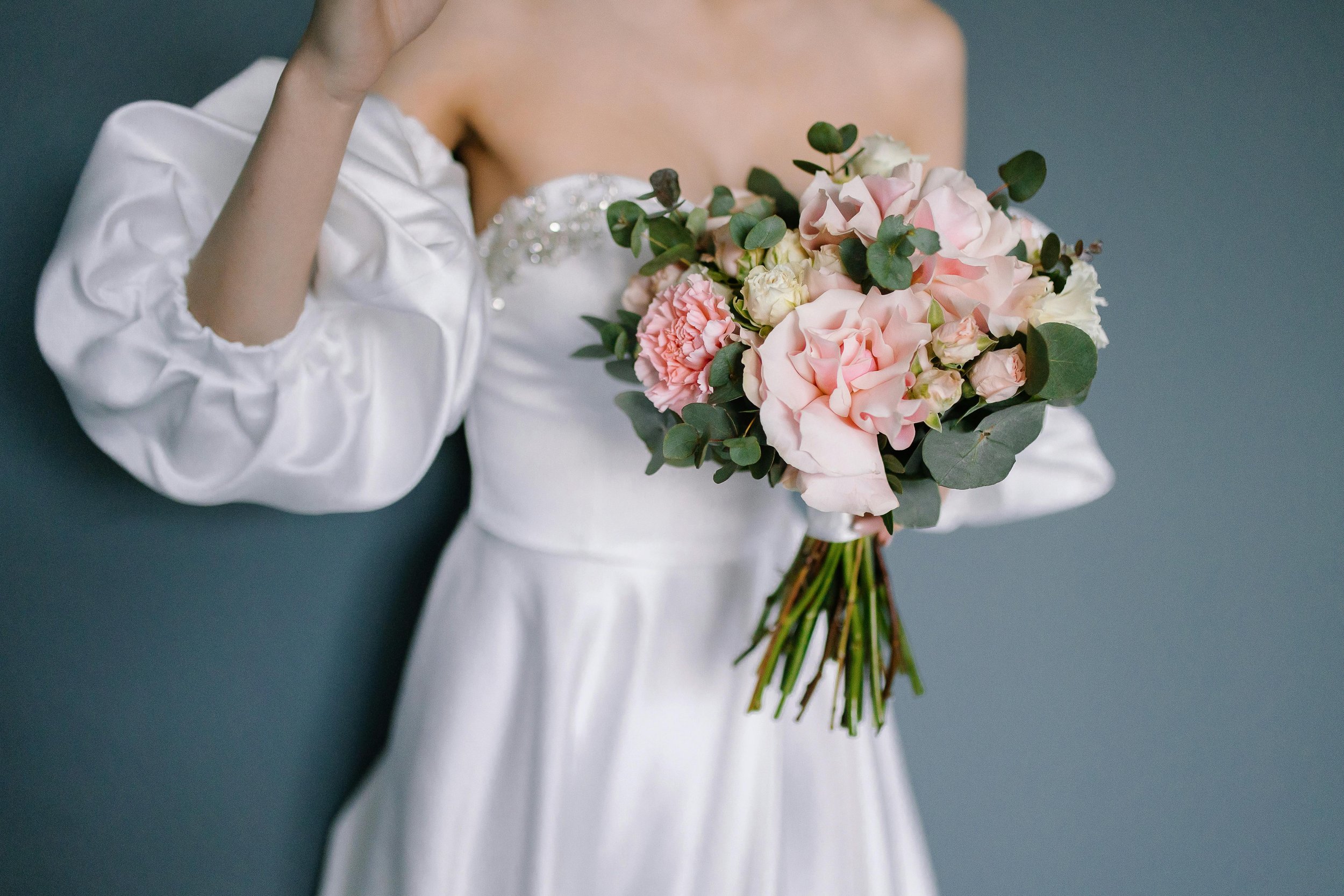 A person in a white wedding dress holding a bouquet of pink and white flowers with green leaves.