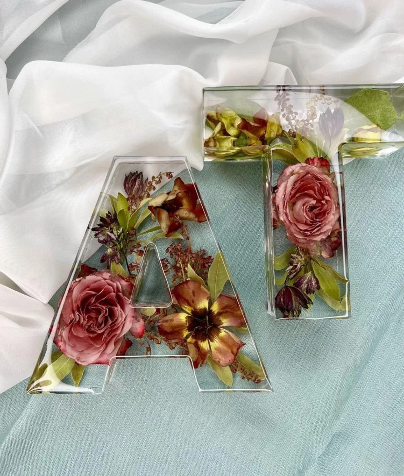 Decorative glass letters 'A' and 'T' filled with pink and burgundy flowers and green leaves, placed on a light blue tablecloth with white fabric in the background.